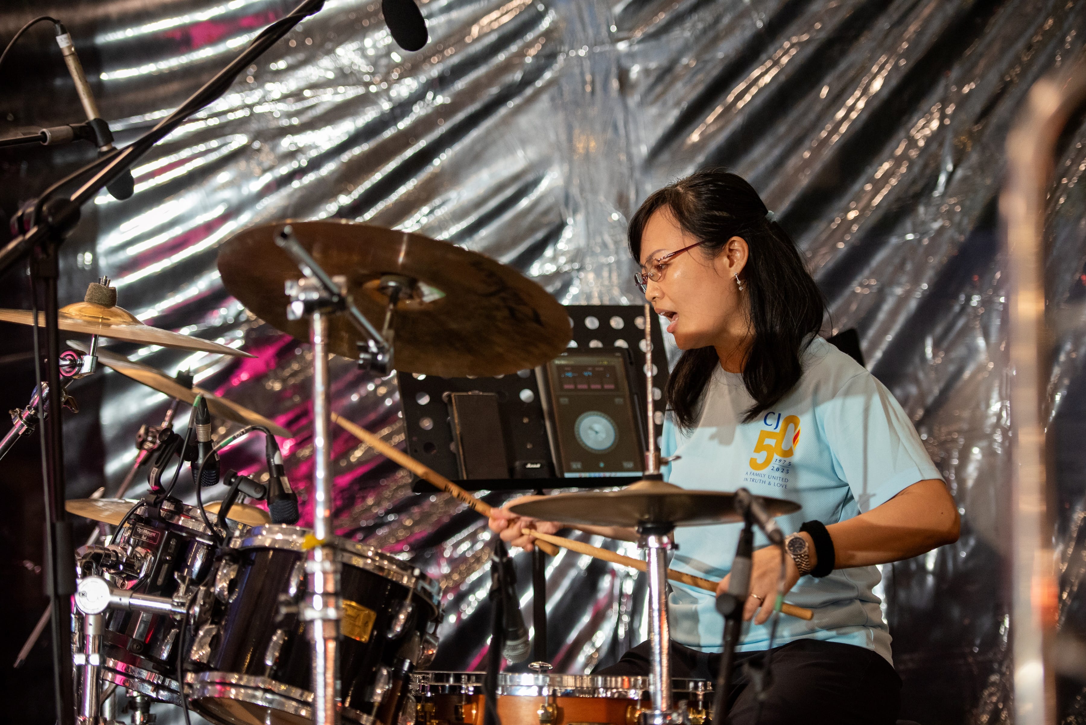 Woman with glasses in a light blue shirt playing drums on stage with cymbals and drumsticks visible.