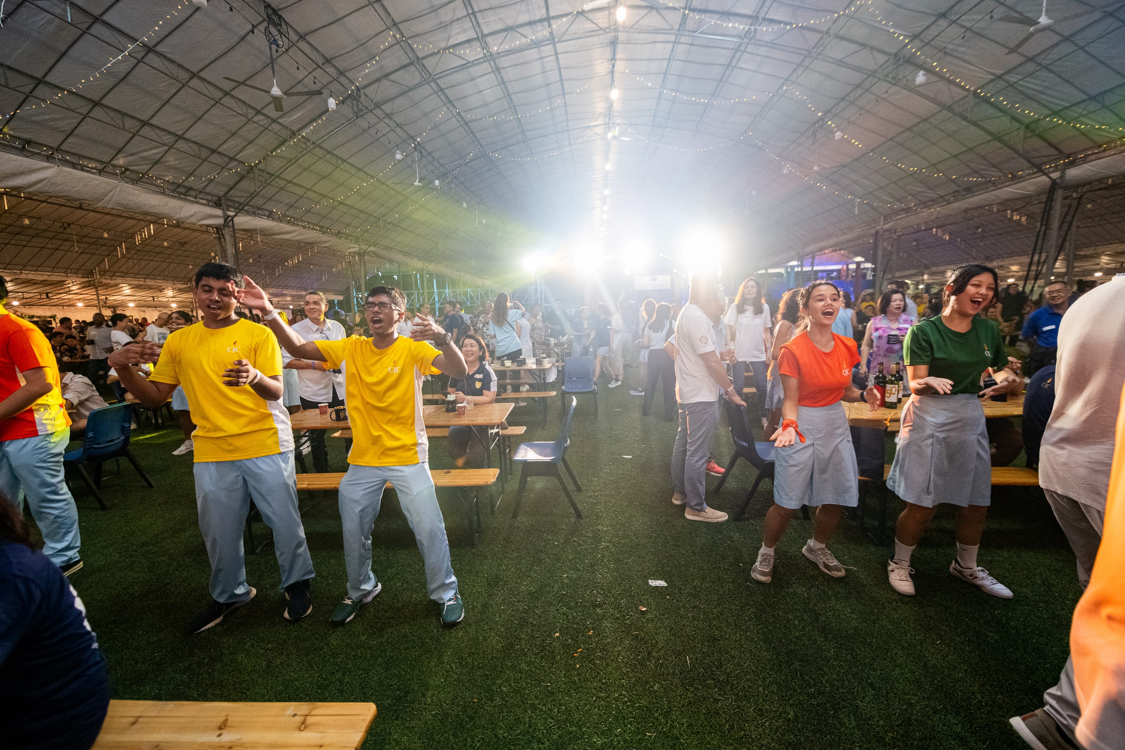 People dance and sing beside long tables under a tent roof with string lights and bright stage lights.