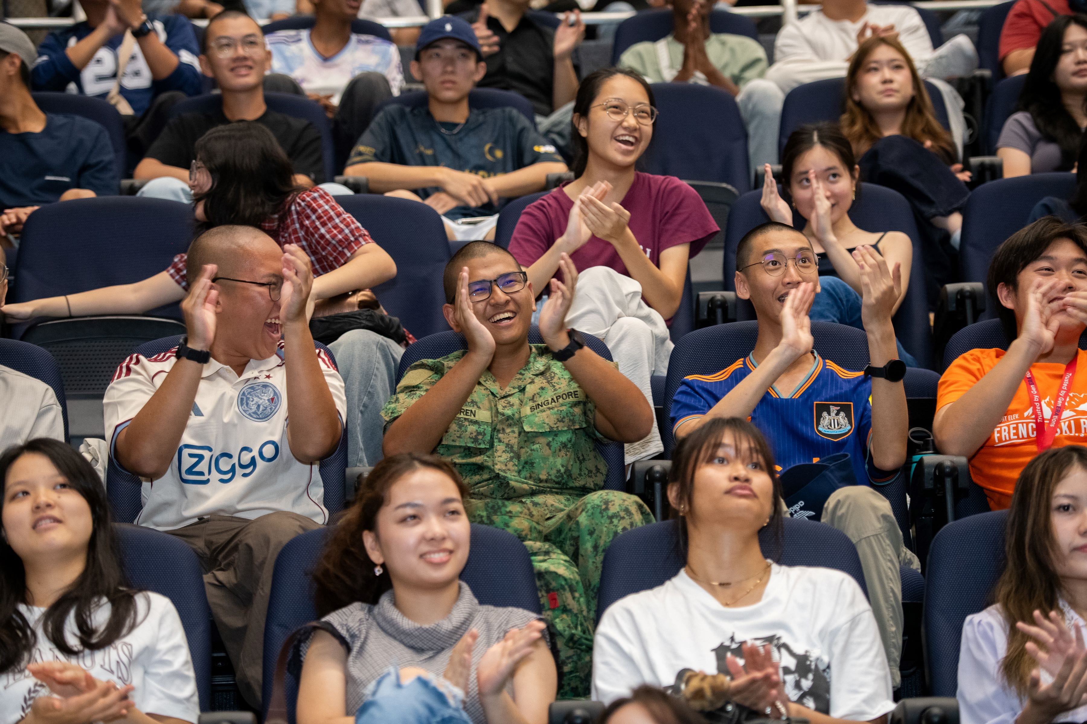 A crowd of smiling people clapping in an auditorium, including one person in a Singapore military uniform.