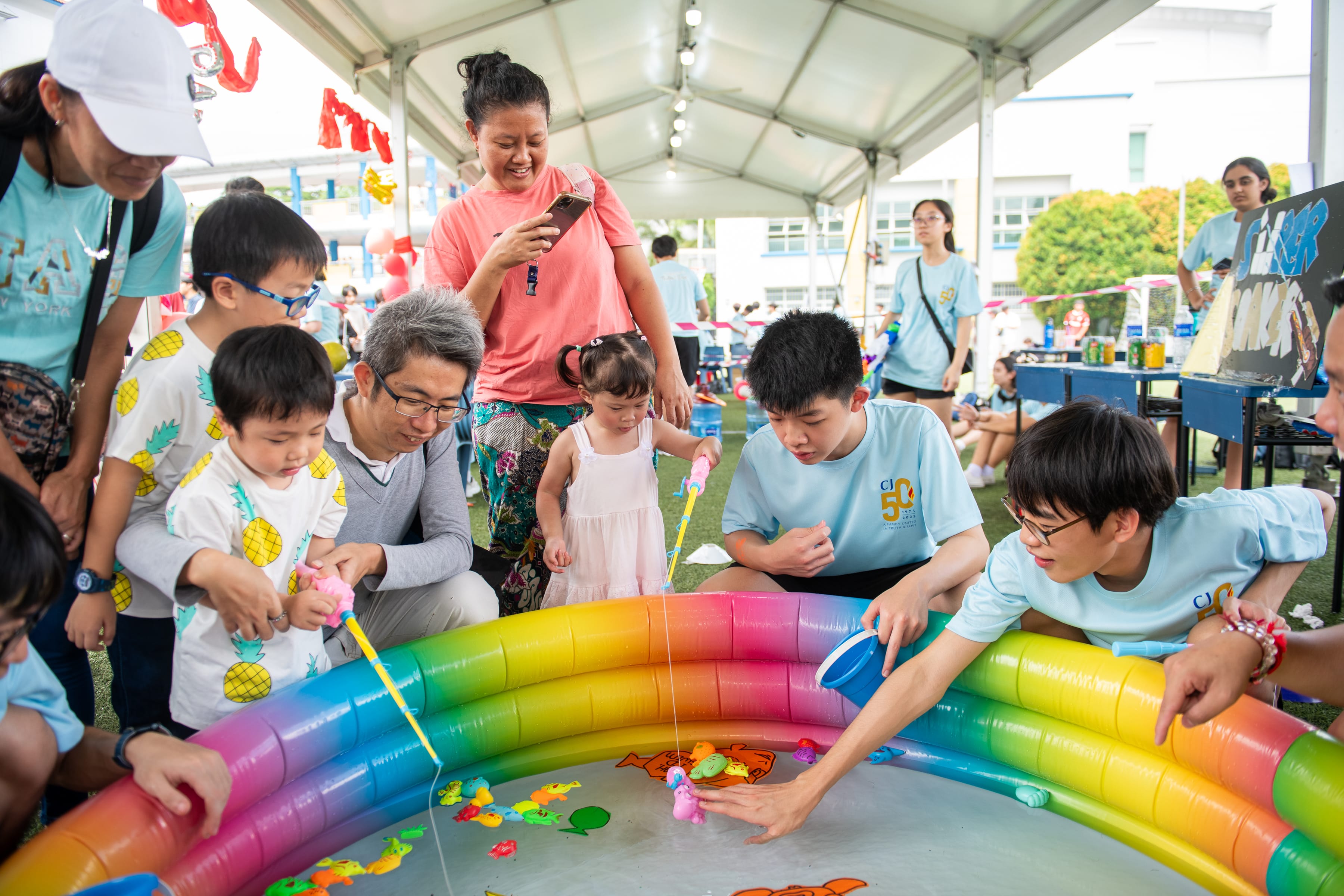 People and kids gather around a rainbow inflatable pool, fishing for toy fish under a canopy.