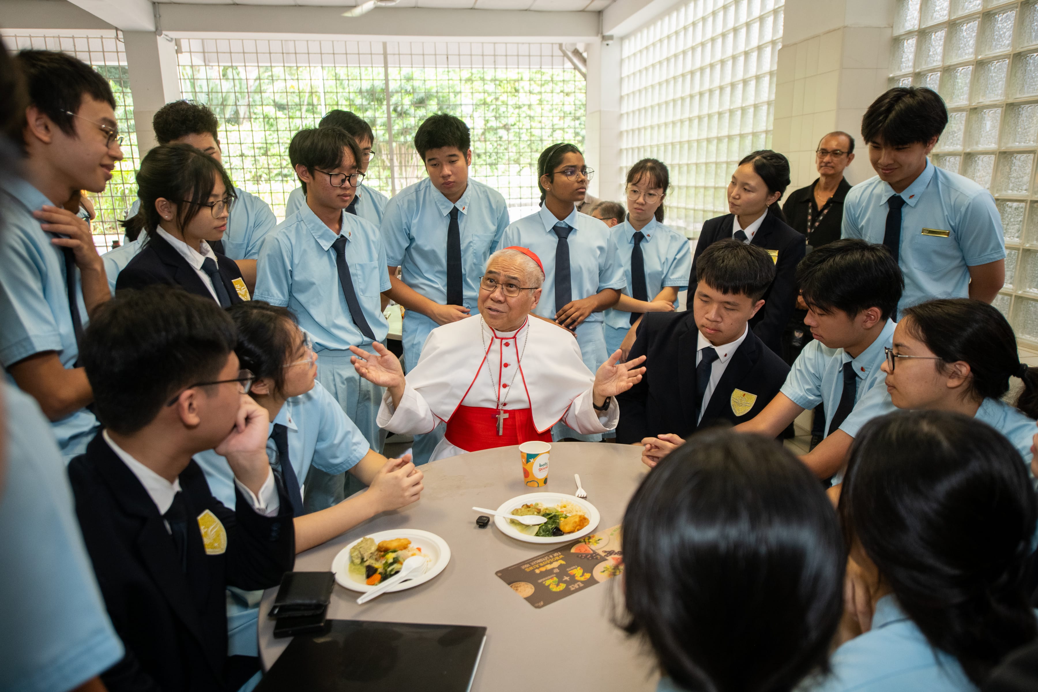 Elderly man in white and red clerical robes speaks at a cafeteria table surrounded by students in uniforms.