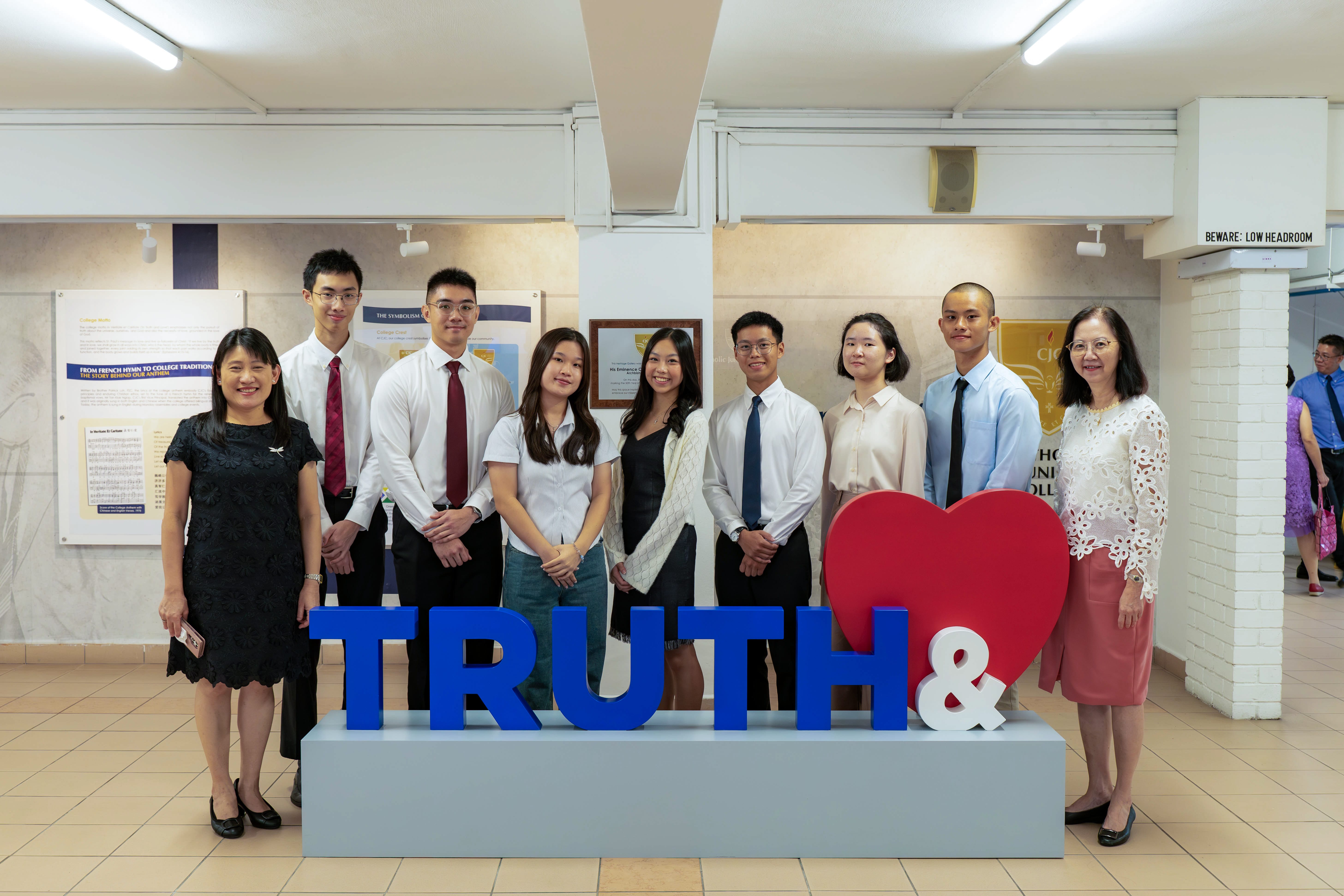 Nine people posing behind a display with blue letters spelling TRUTH and a red heart symbol in an indoor hallway.