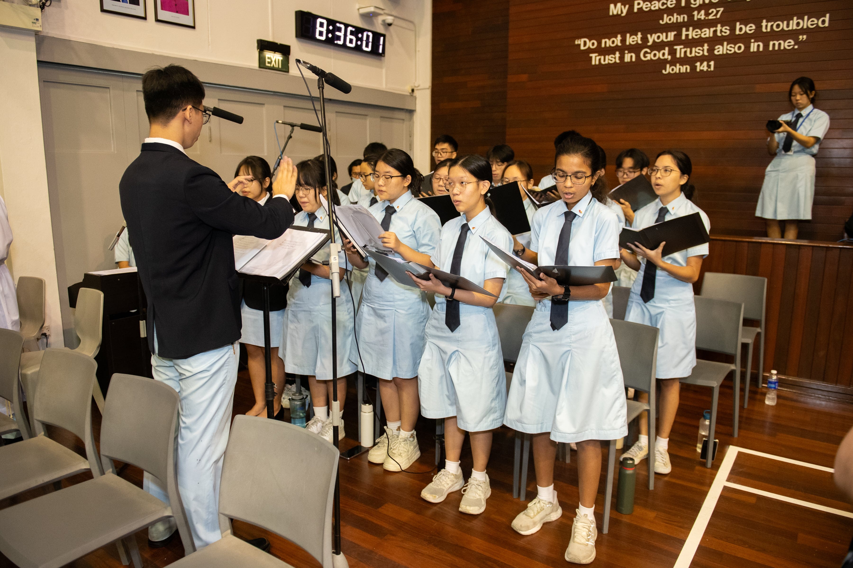 Students in light blue uniforms sing in a choir led by a conductor in a chapel with a Bible verse on the wall.