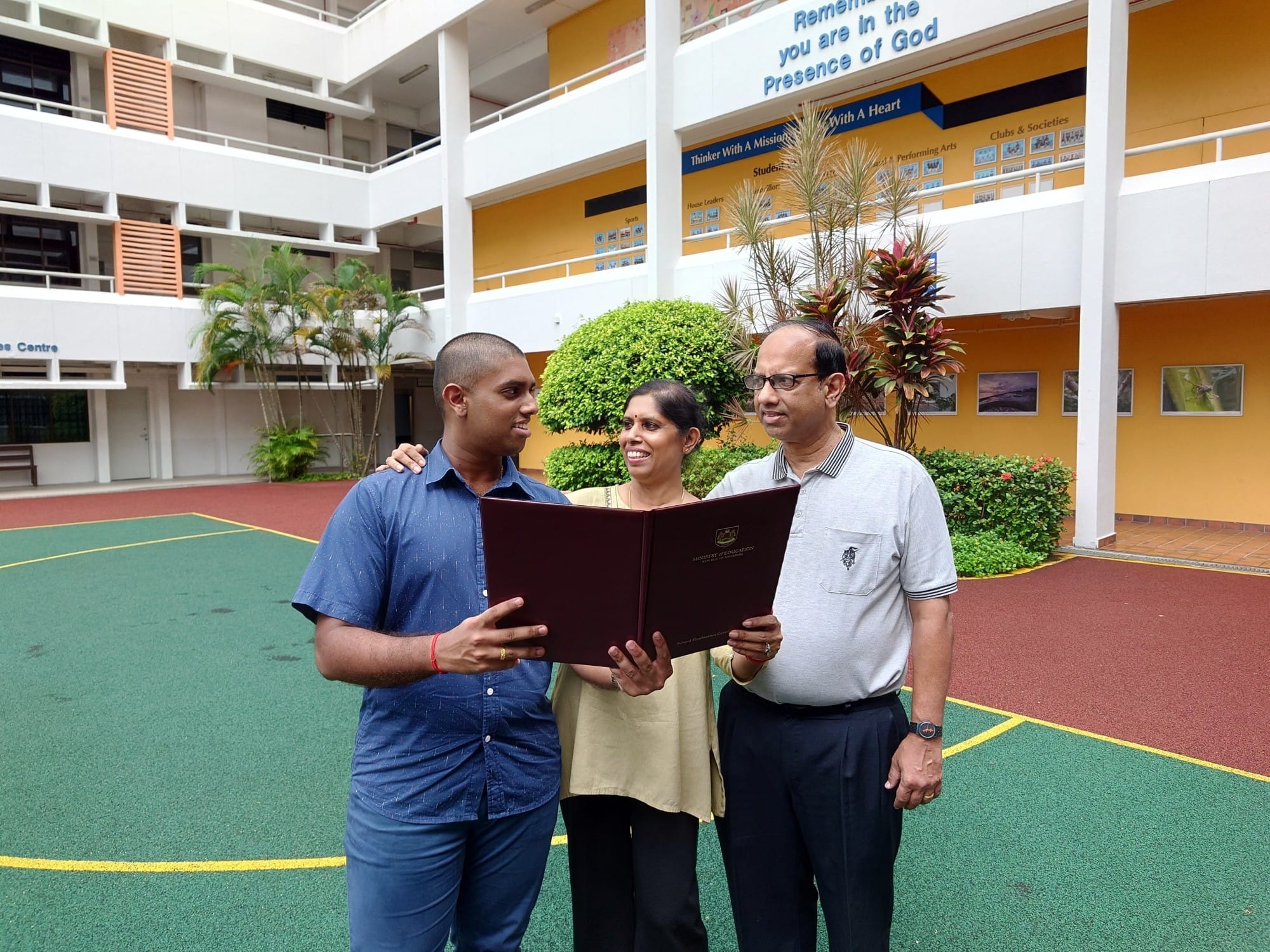 Three people stand in a school courtyard, looking at an open certificate folder together.