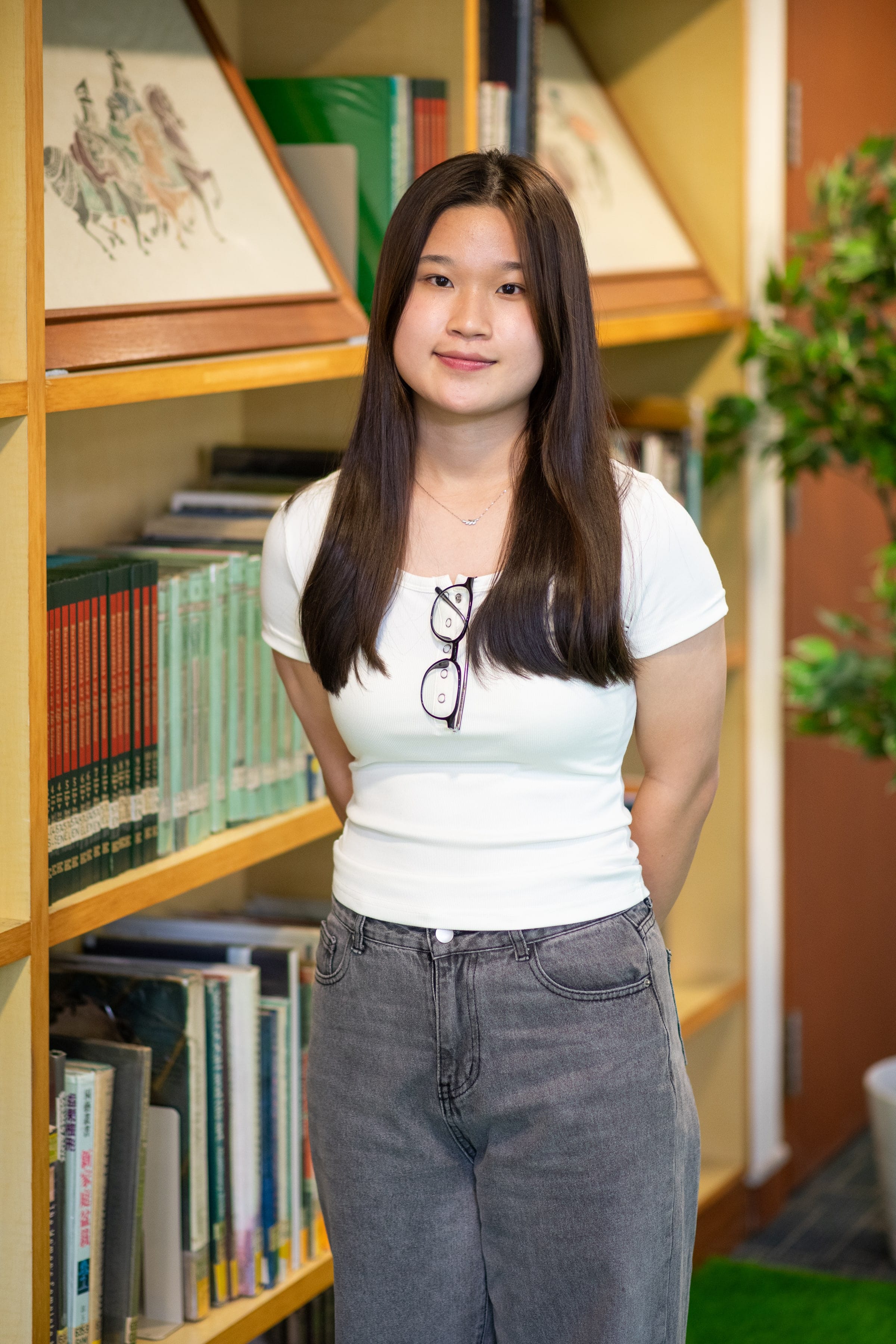 Young woman with long dark hair, white t-shirt, gray jeans, glasses tucked in collar, standing before a bookshelf.