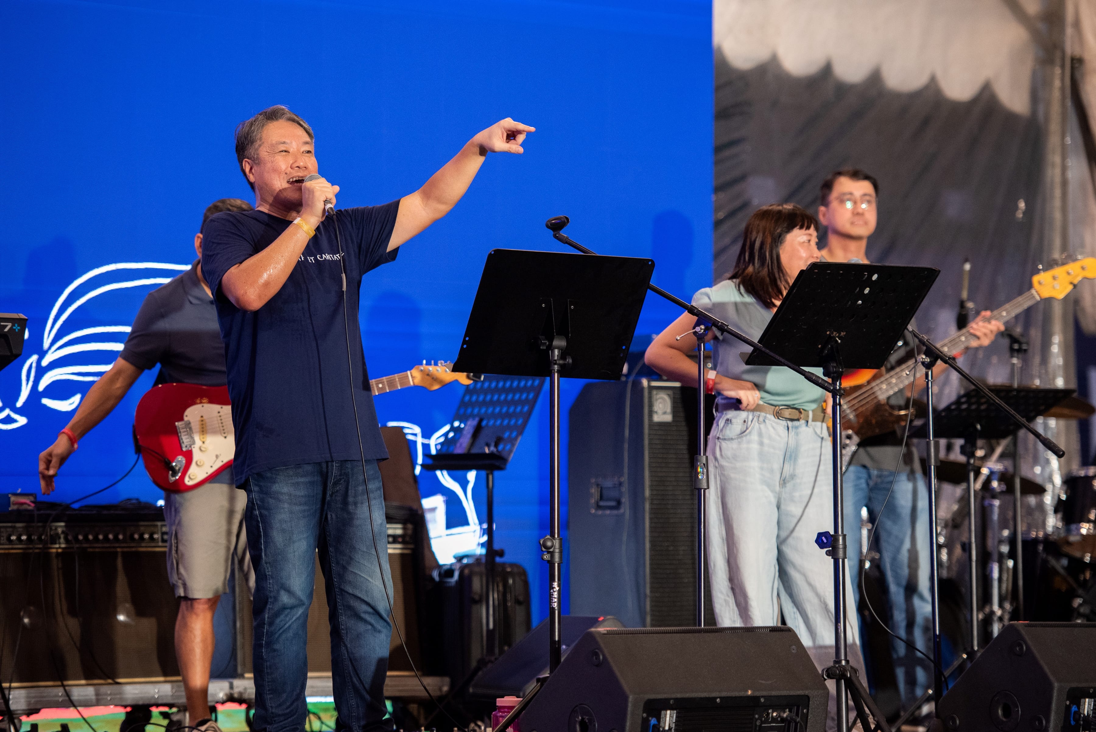Man sings into a microphone and points on stage while bandmates play guitar and bass behind music stands.