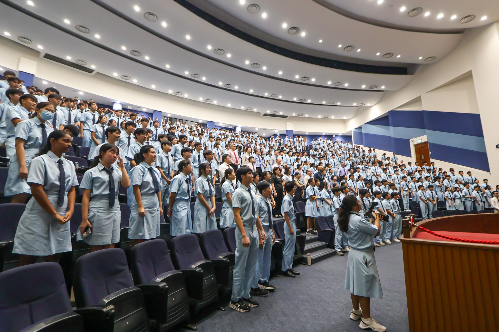 Hundreds of students in light blue uniforms stand in a large auditorium during a school assembly.