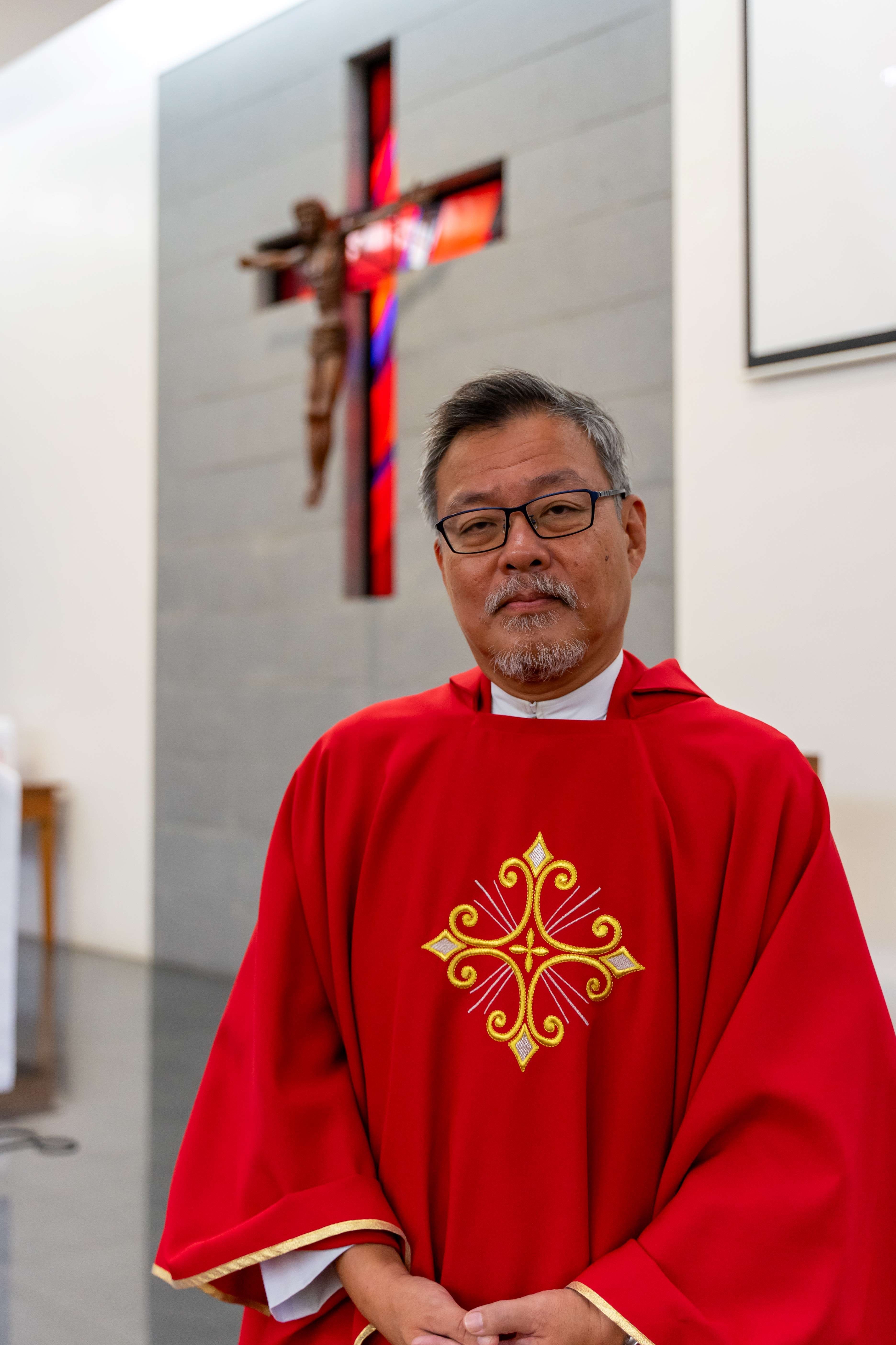 A middle-aged man in red vestments with gold embroidery stands before a crucifix on a church wall.
