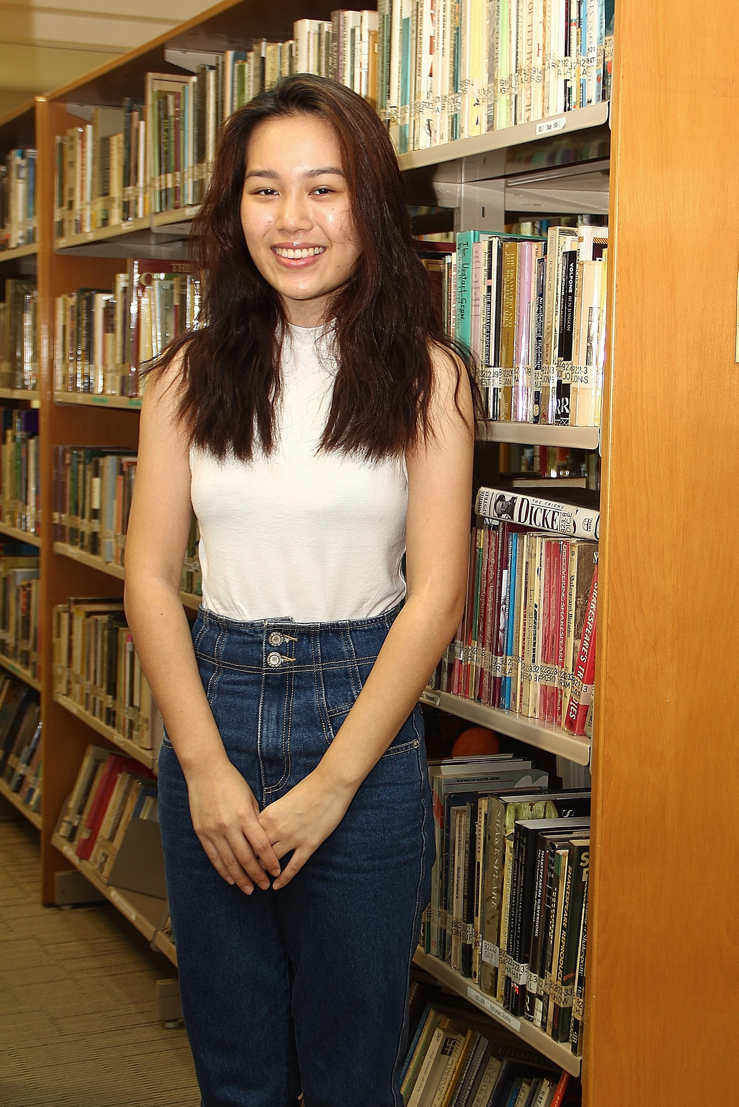 Smiling woman in a white sleeveless top and blue jeans stands between library bookshelves.