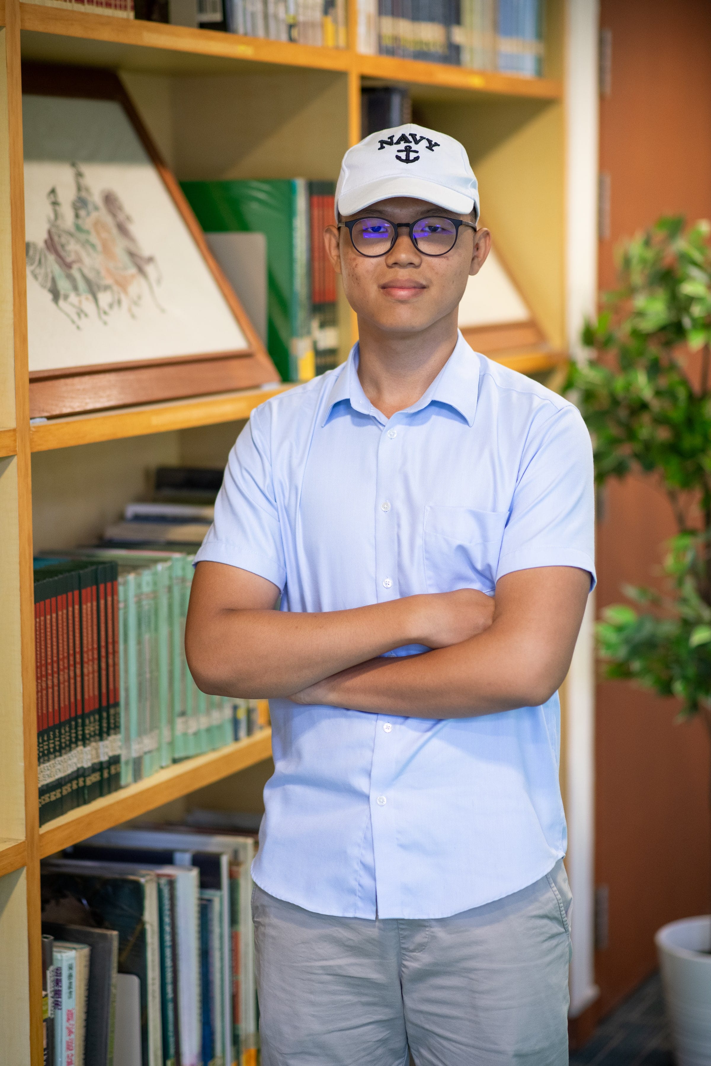 Young man with glasses and a white Navy cap, arms crossed, wearing a light blue shirt, standing in front of a bookshelf.