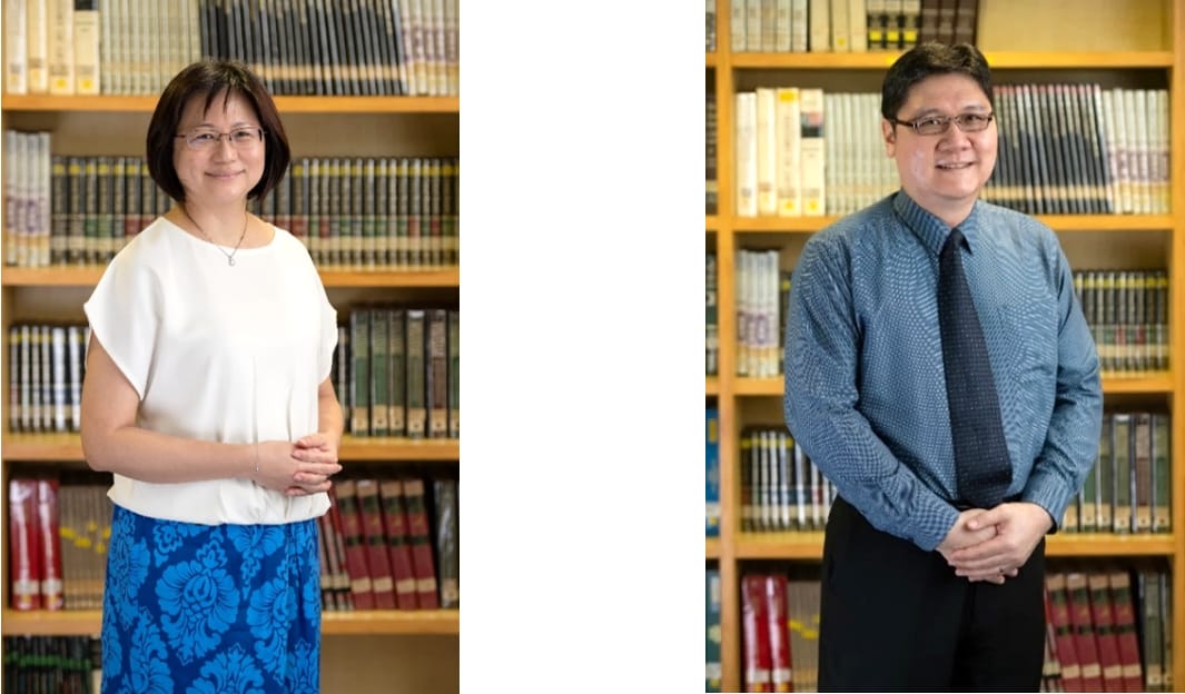 Two people wearing glasses stand smiling in front of wooden bookshelves filled with books.