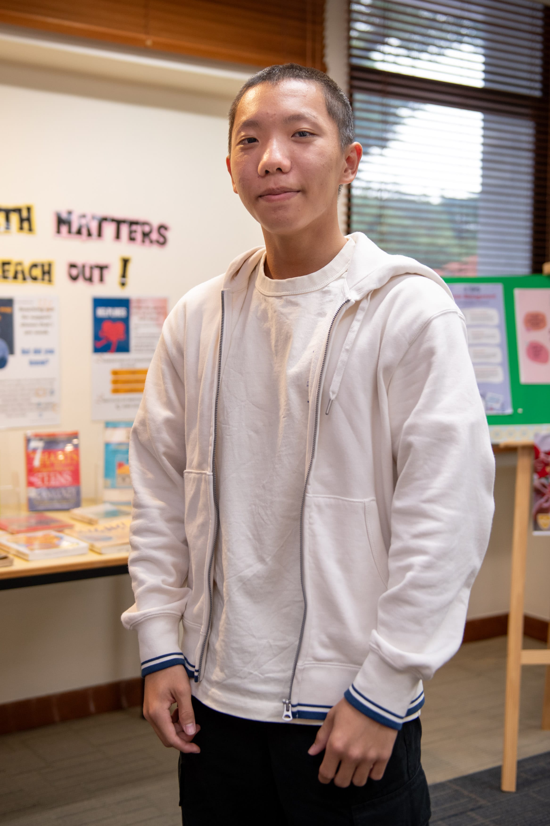 Young man with short hair wearing a white zip-up hoodie, standing in a library with a bulletin board behind him.