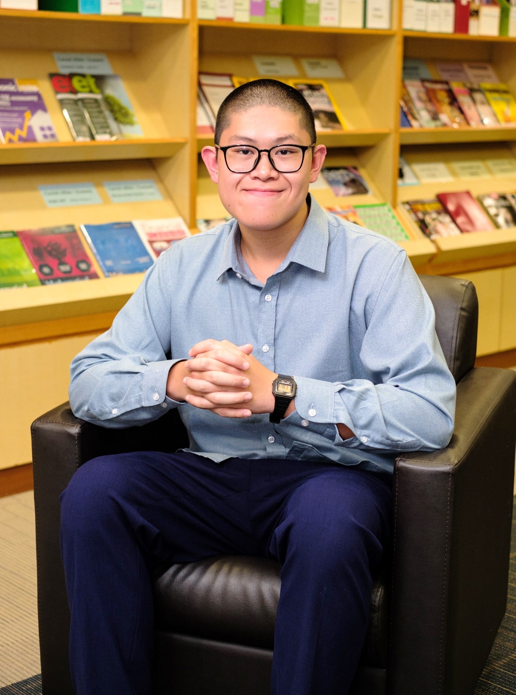 Young man with glasses and a watch sits in a dark leather chair, smiling, in front of shelves of magazines.