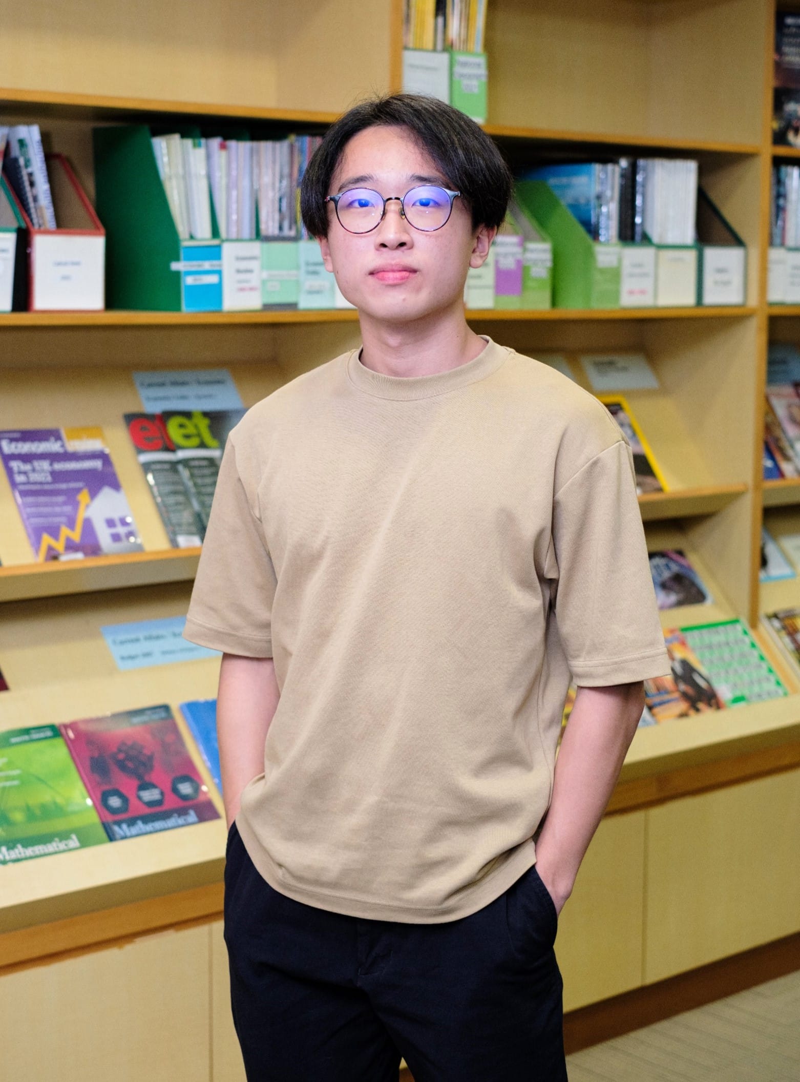 Young man with glasses and a beige t-shirt standing in front of wooden shelves filled with books and magazines.