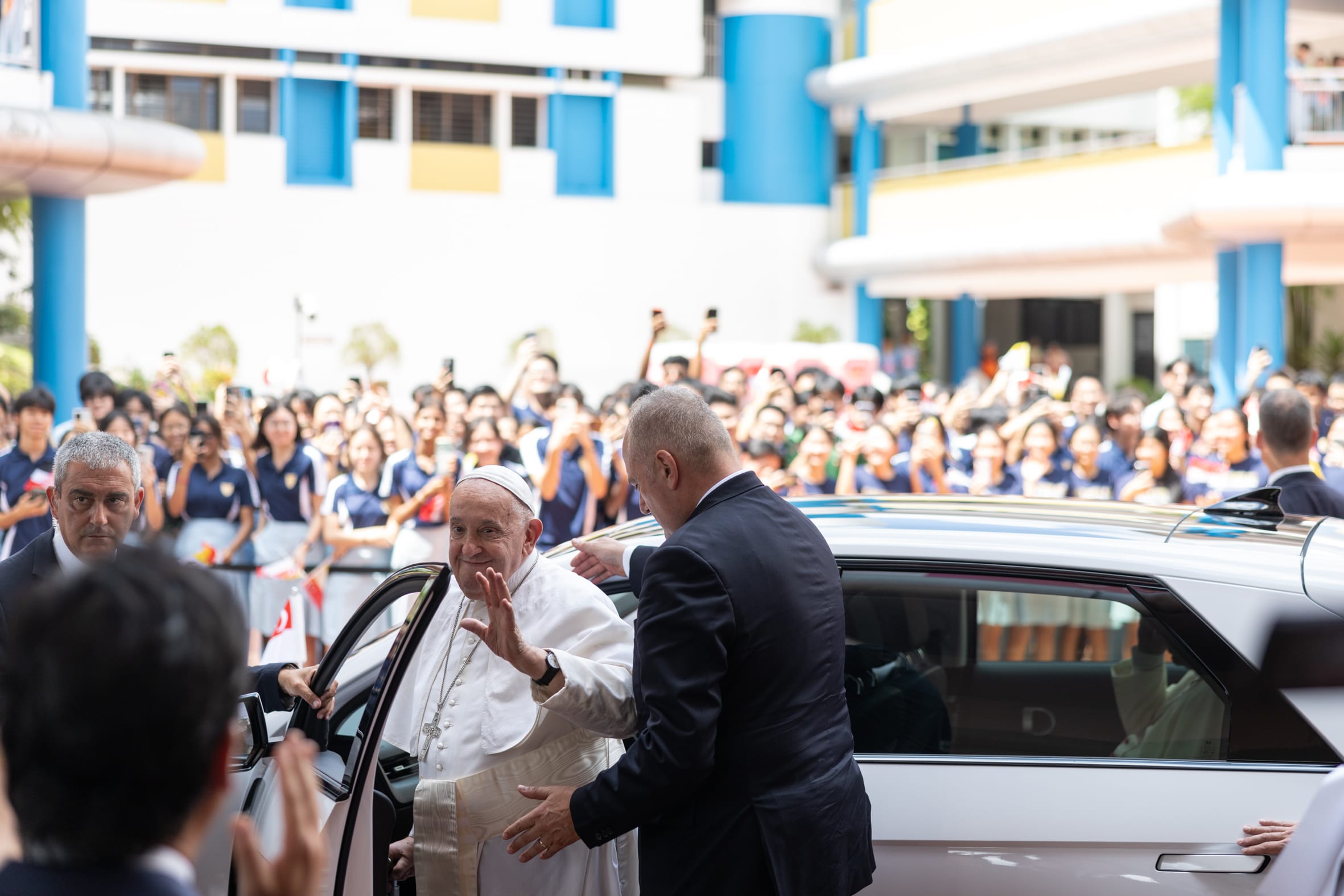 An elderly man in white papal robes waves while entering a car, surrounded by a large crowd of cheering students.