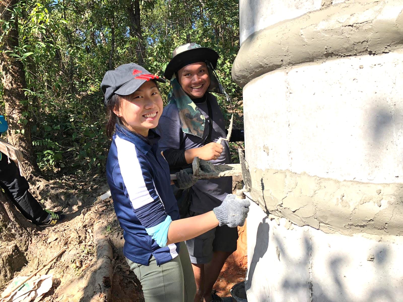Two smiling people wearing caps and gloves apply cement to a concrete structure outdoors surrounded by trees.