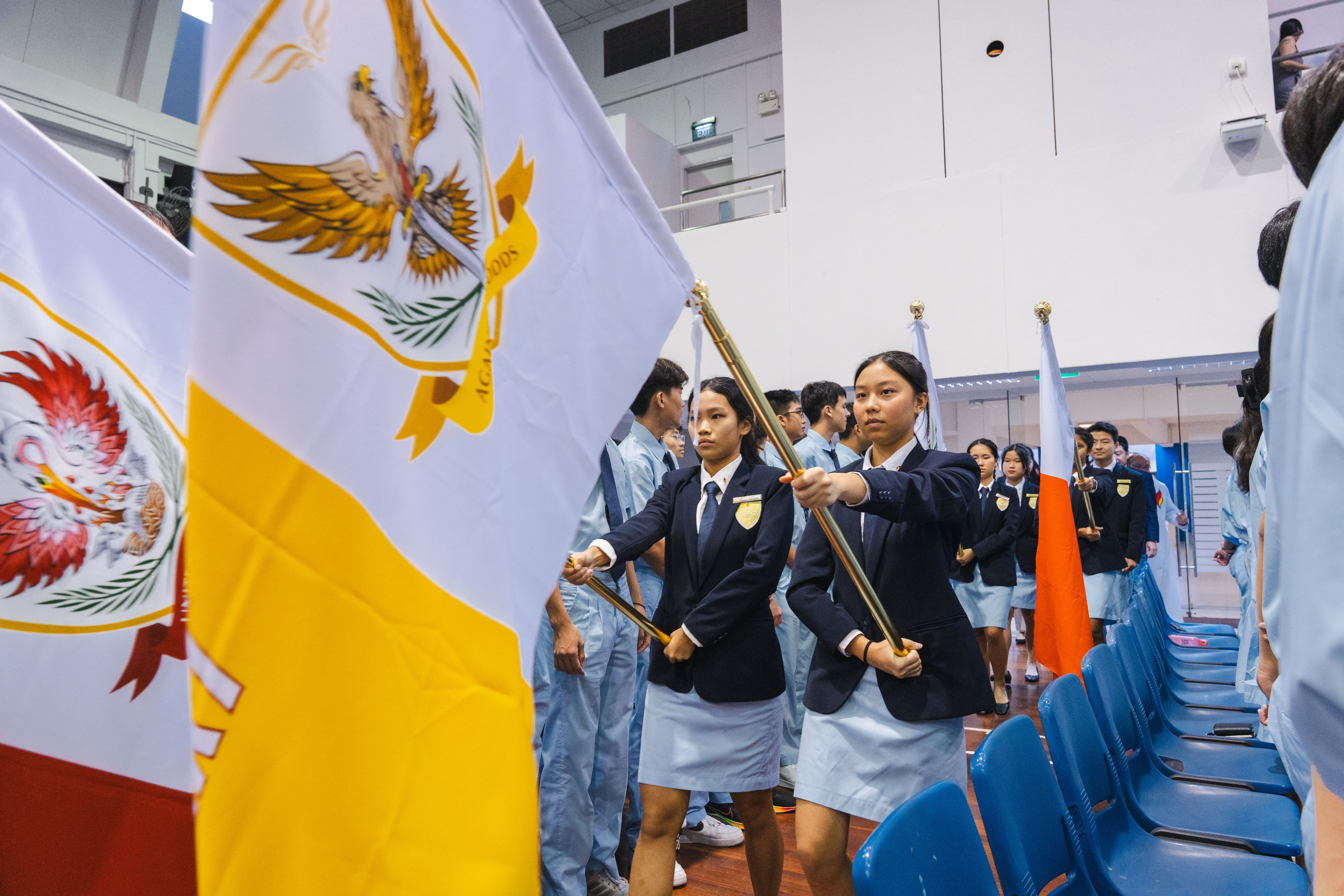 Students in uniforms march indoors carrying flags; a large white-and-yellow flag fills the left foreground.
