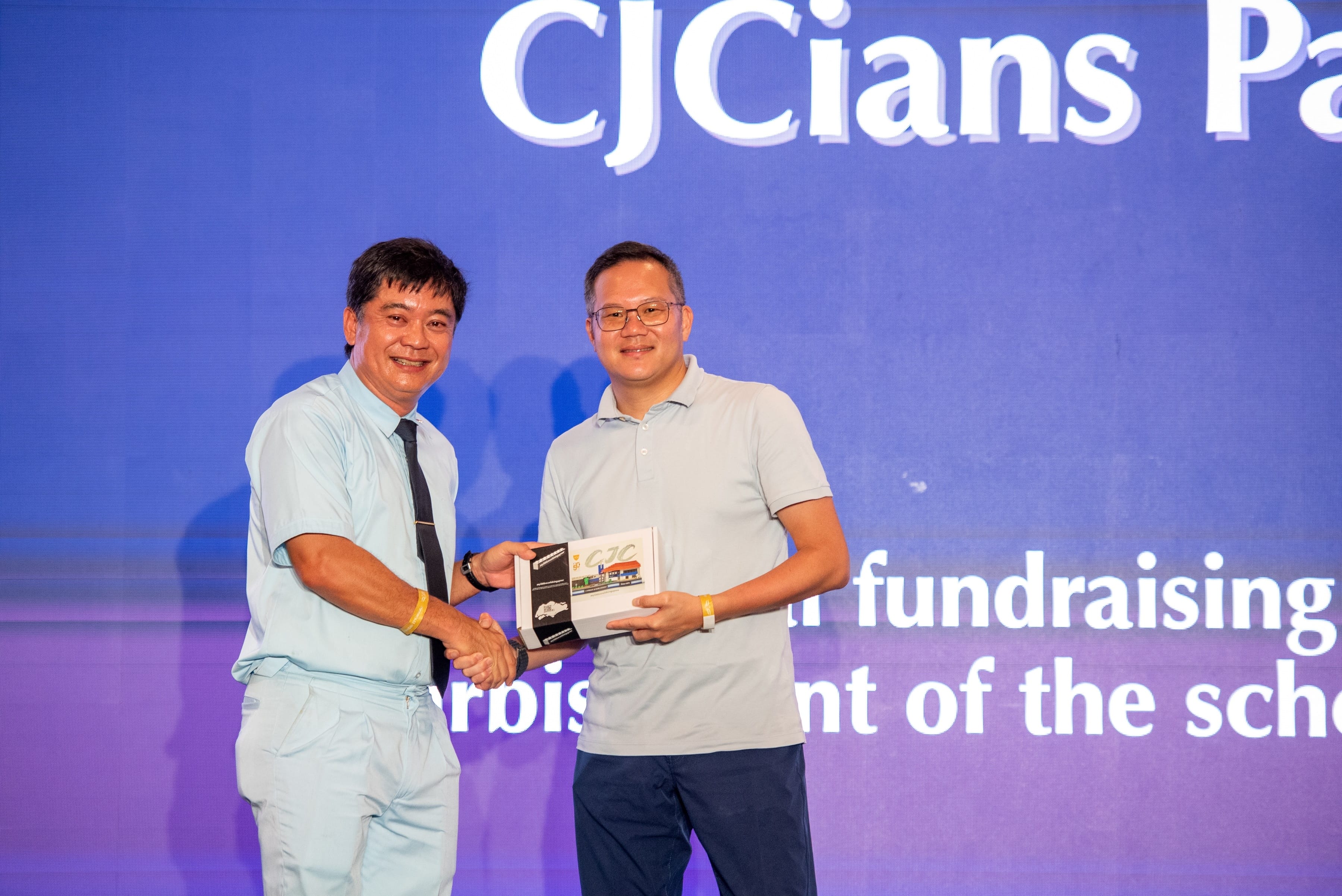 Two men smile and shake hands while holding a plaque on a stage with a blue CJCians backdrop.