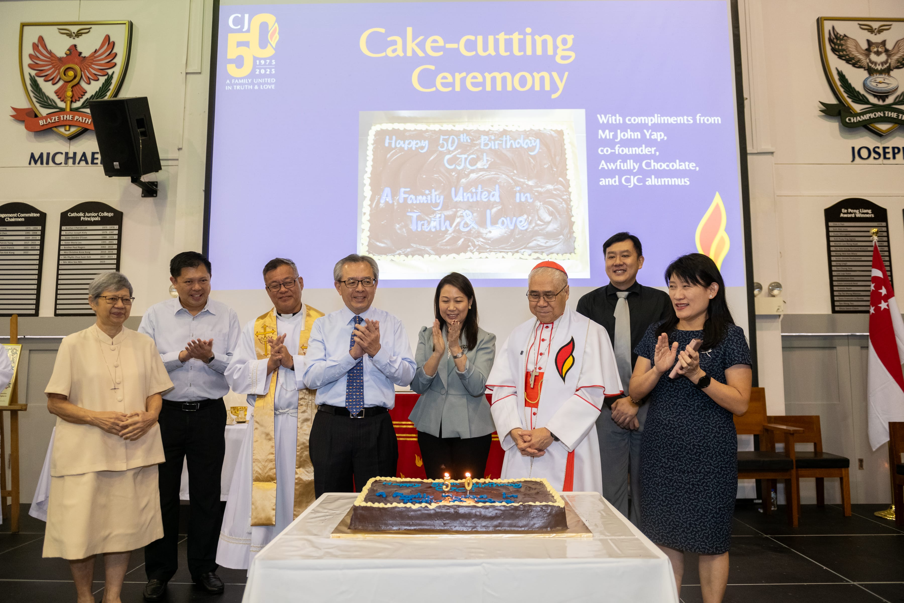 Group applauds around a cake with 50 candles; screen behind reads Cake-cutting Ceremony and Happy 50th Birthday.