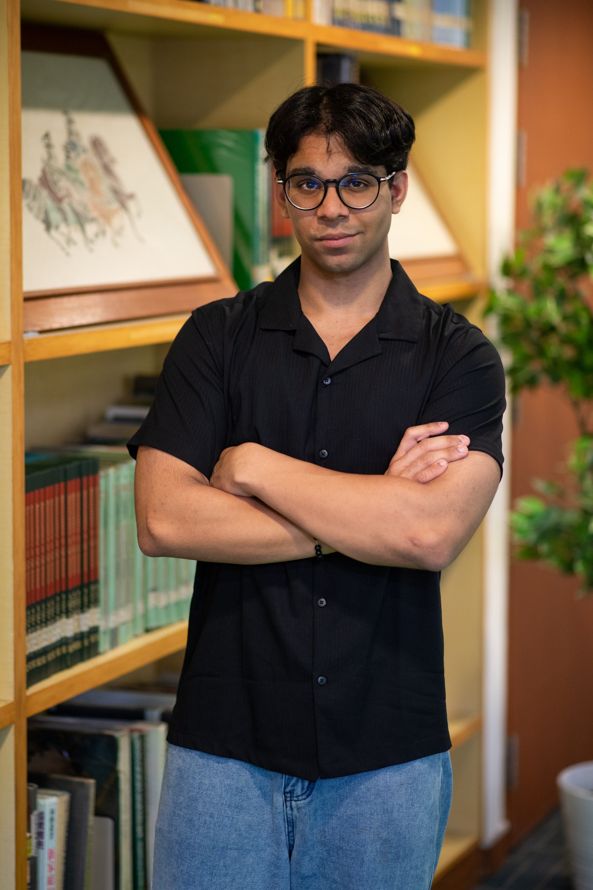 Young man with glasses and black shirt stands arms crossed before a wooden bookshelf filled with books.