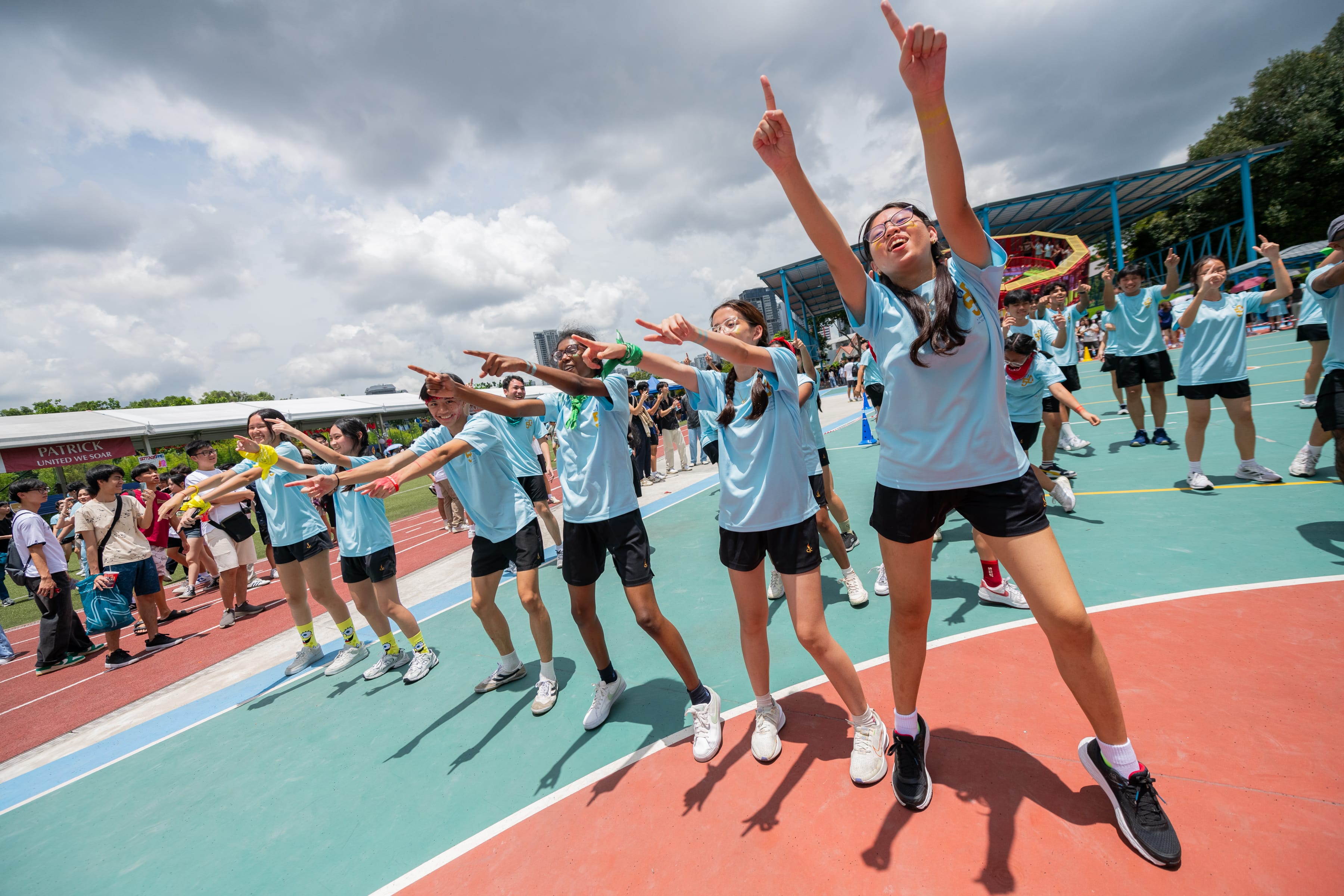 Students in light blue shirts cheer and point on a track, with a crowd and cloudy sky behind.