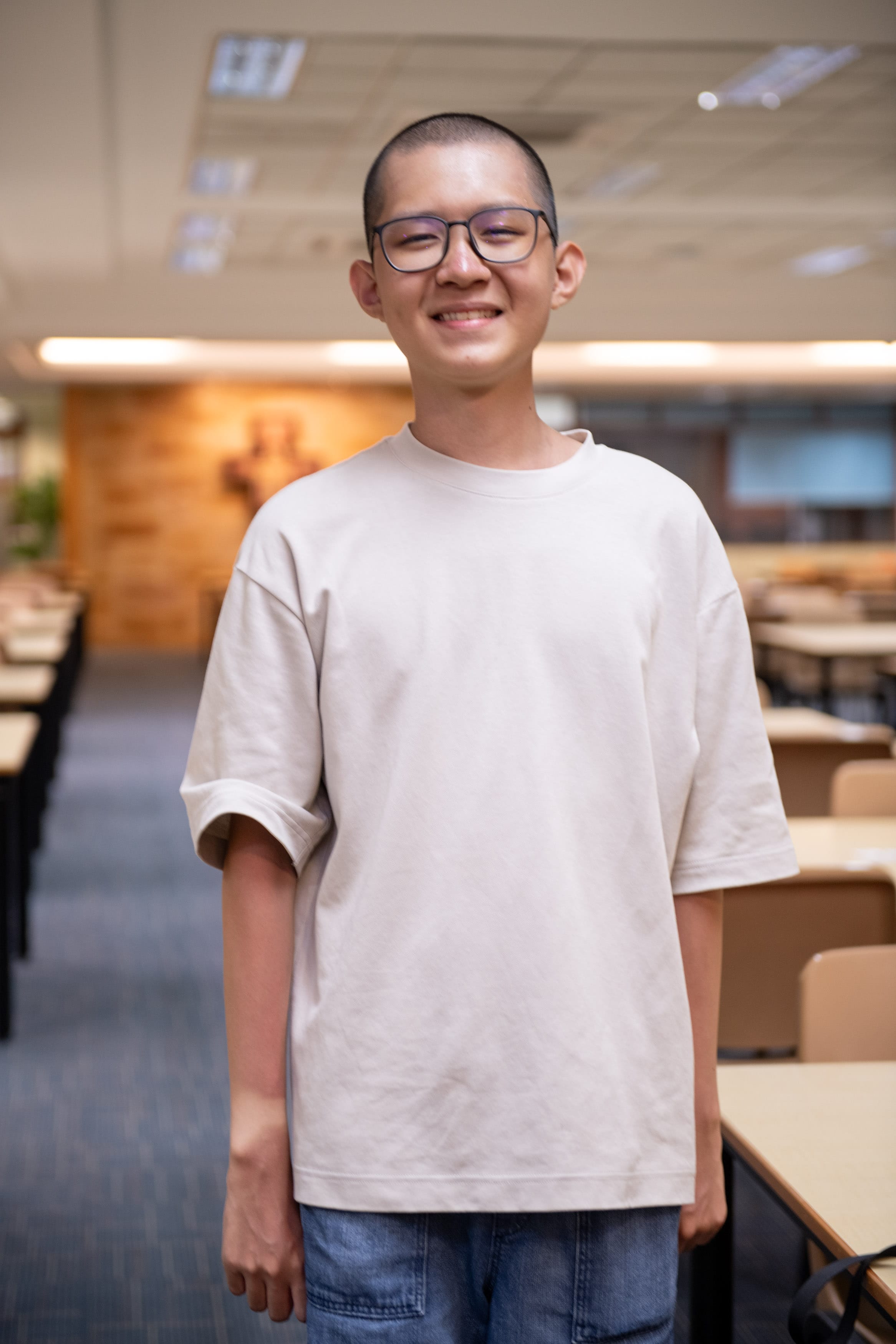 Young man with shaved head and glasses smiling, wearing a beige oversized t-shirt, standing in a classroom.