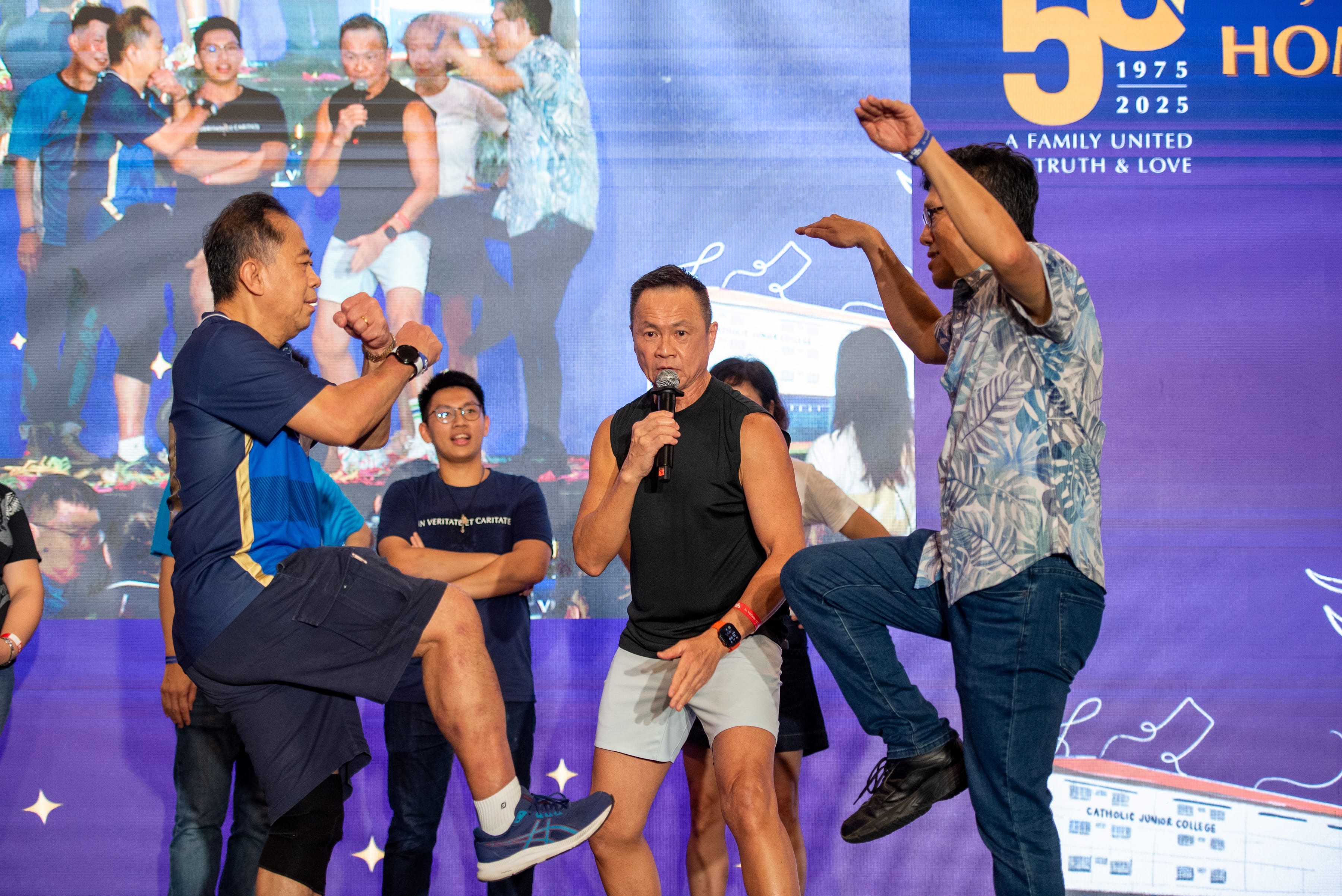 Three men on stage perform playful martial arts poses, one holding a microphone, at a Catholic Junior College 50th anniversary event.