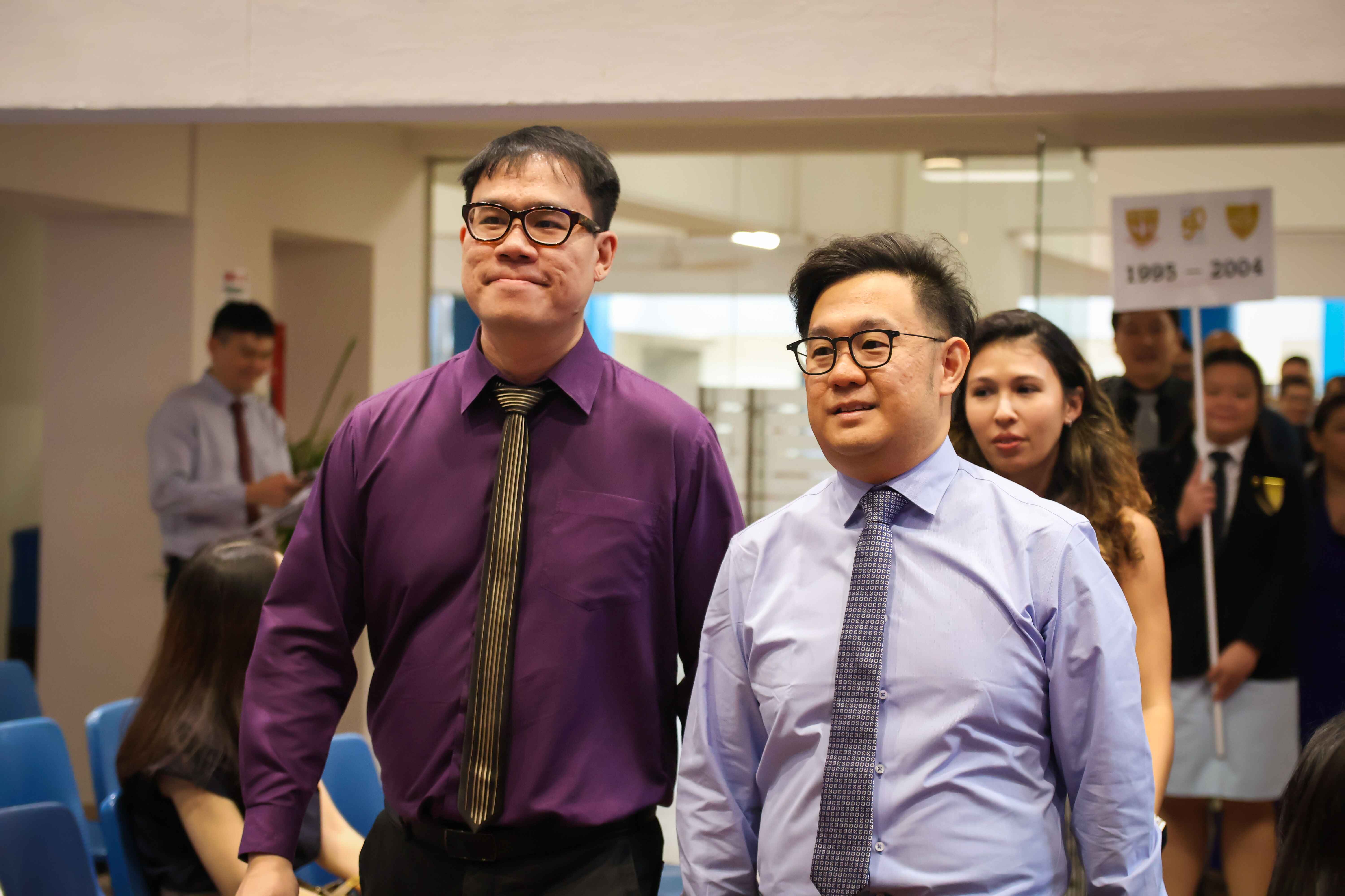 Two men wearing glasses and ties, one in purple and one in blue shirts, stand together at a formal indoor event.