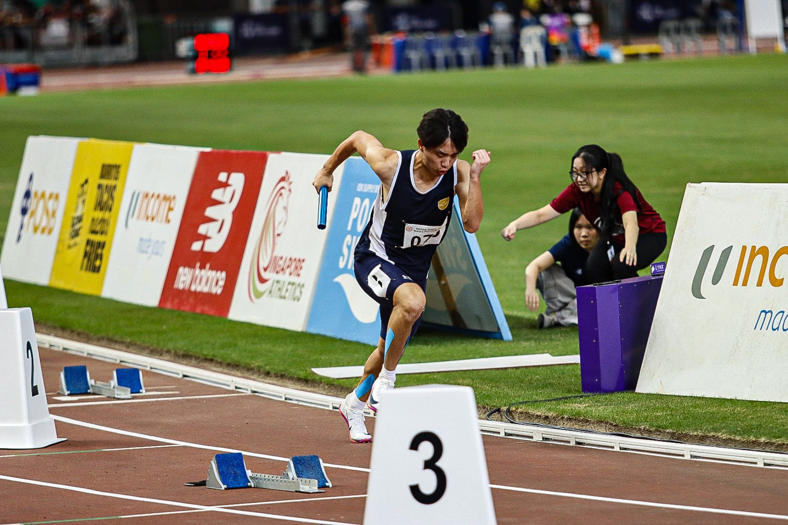 Male relay runner in navy vest sprints from starting blocks on lane 3, holding a blue baton at a track meet.
