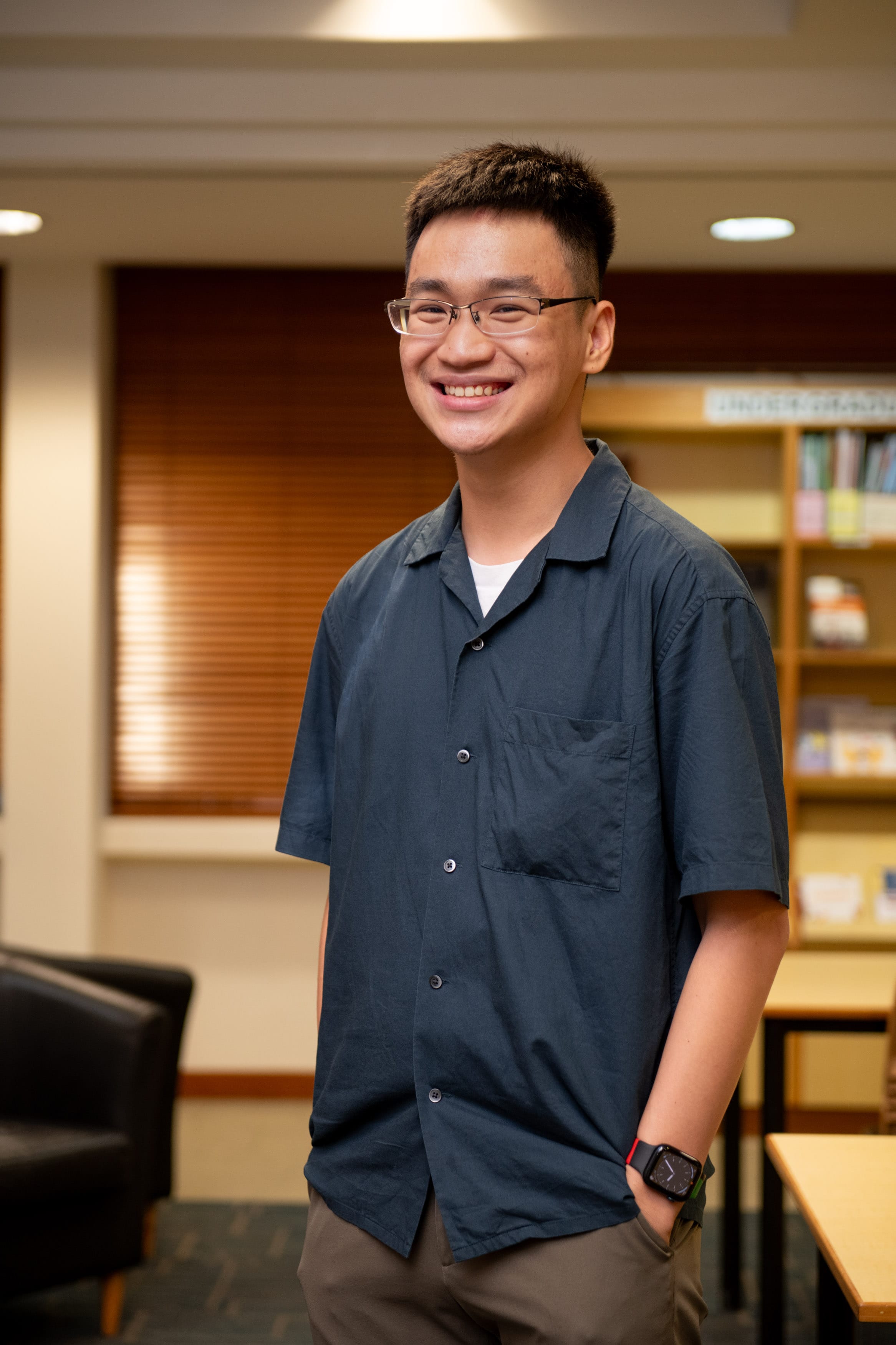 Young man with glasses and a dark blue short-sleeve shirt smiling in a library setting with bookshelves behind him.