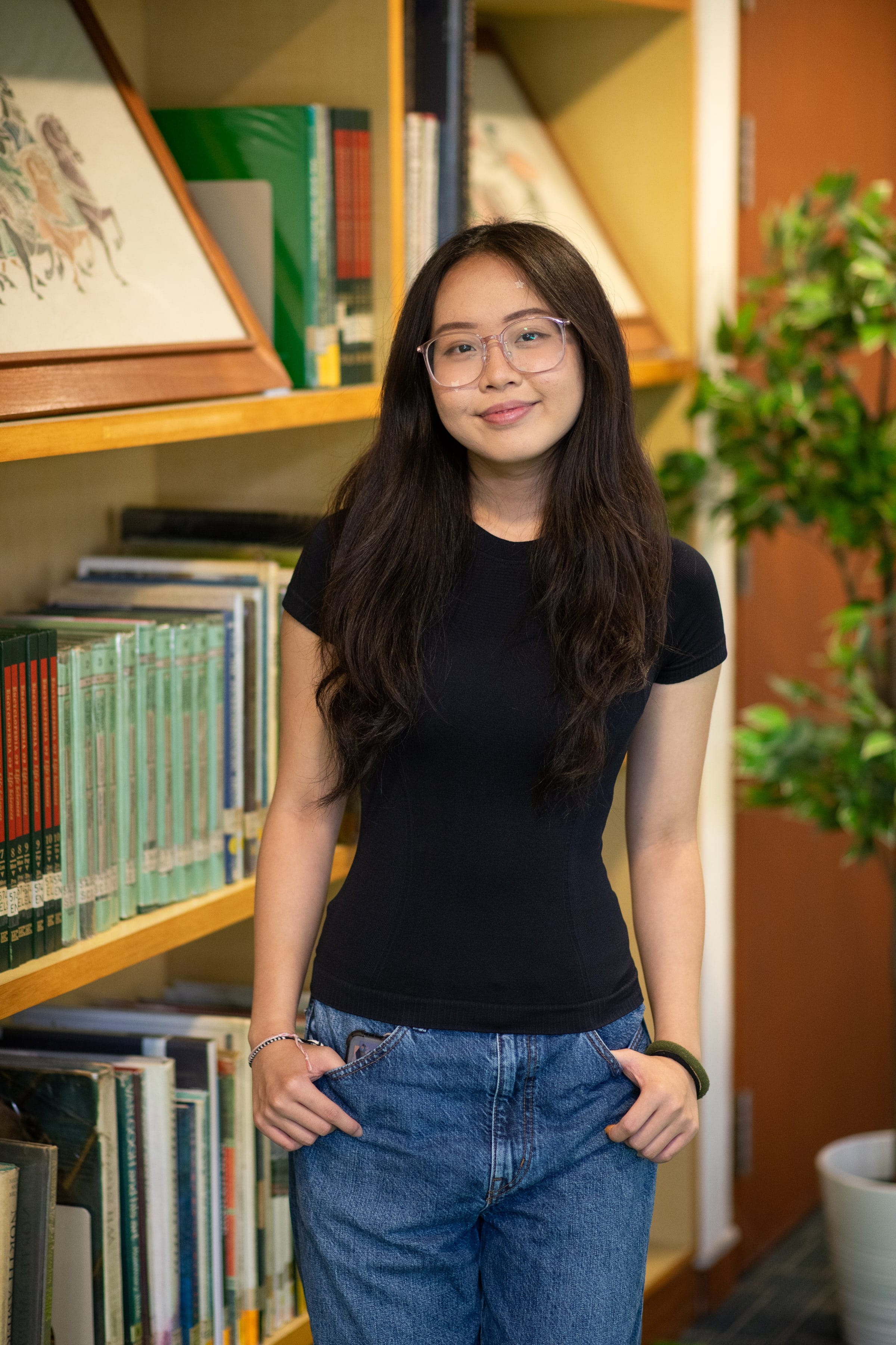 Young woman with long dark hair and clear glasses, wearing a black shirt and jeans, standing in front of bookshelves.