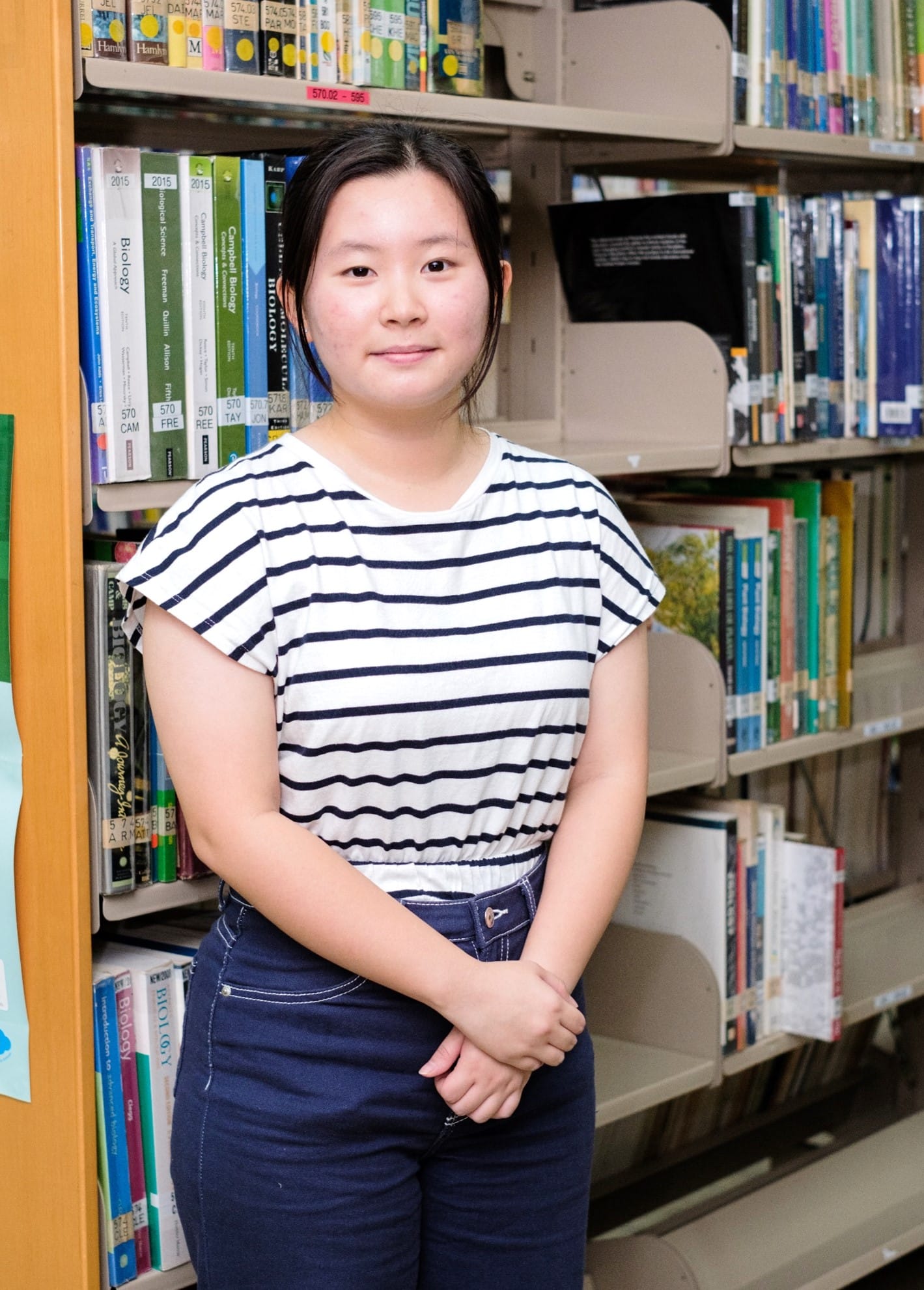 Young woman in a white and navy striped shirt stands smiling in front of library shelves filled with books.