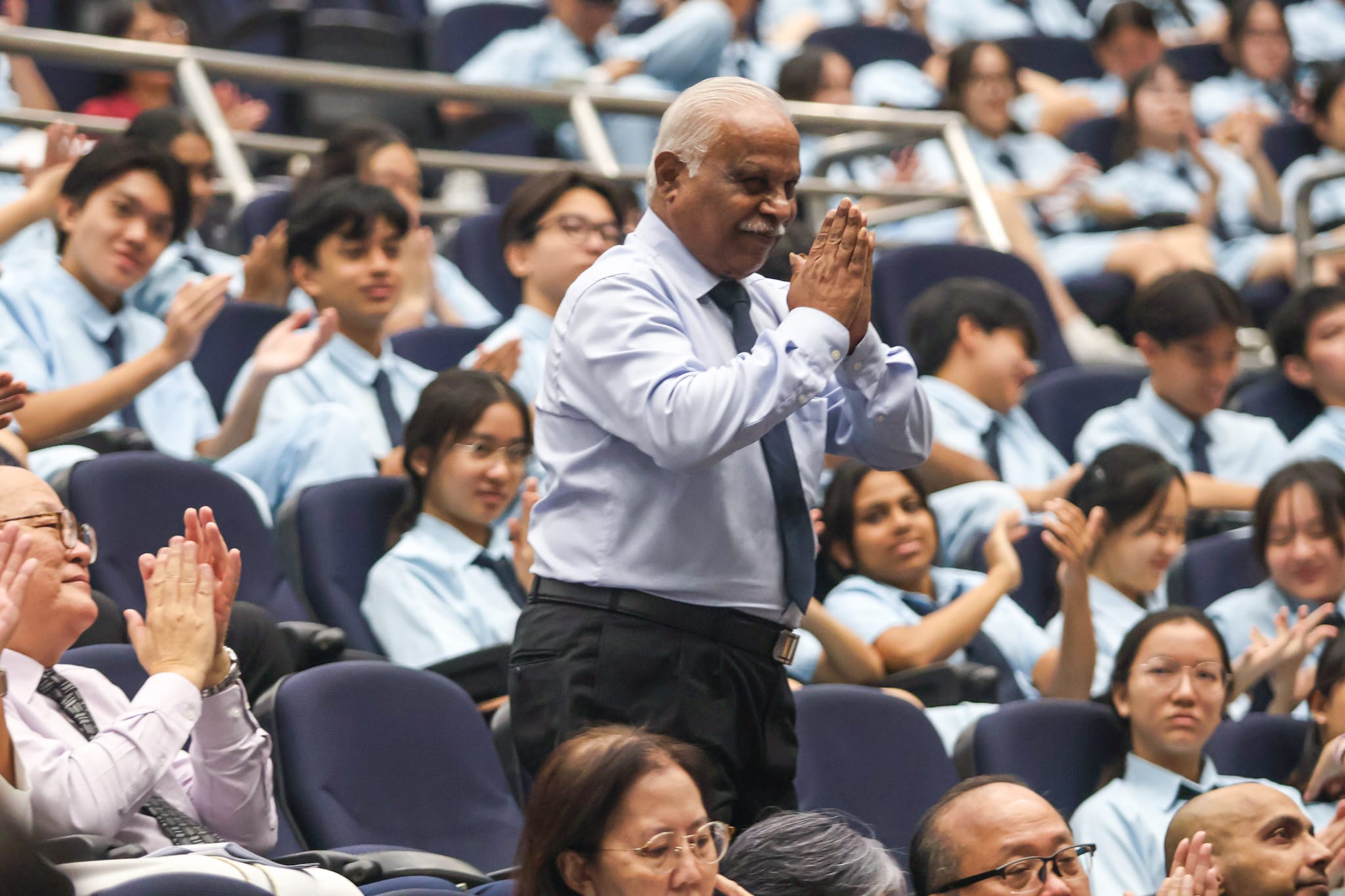An elderly man stands in an auditorium with hands pressed together in a greeting gesture, surrounded by applauding students in light blue uniforms.