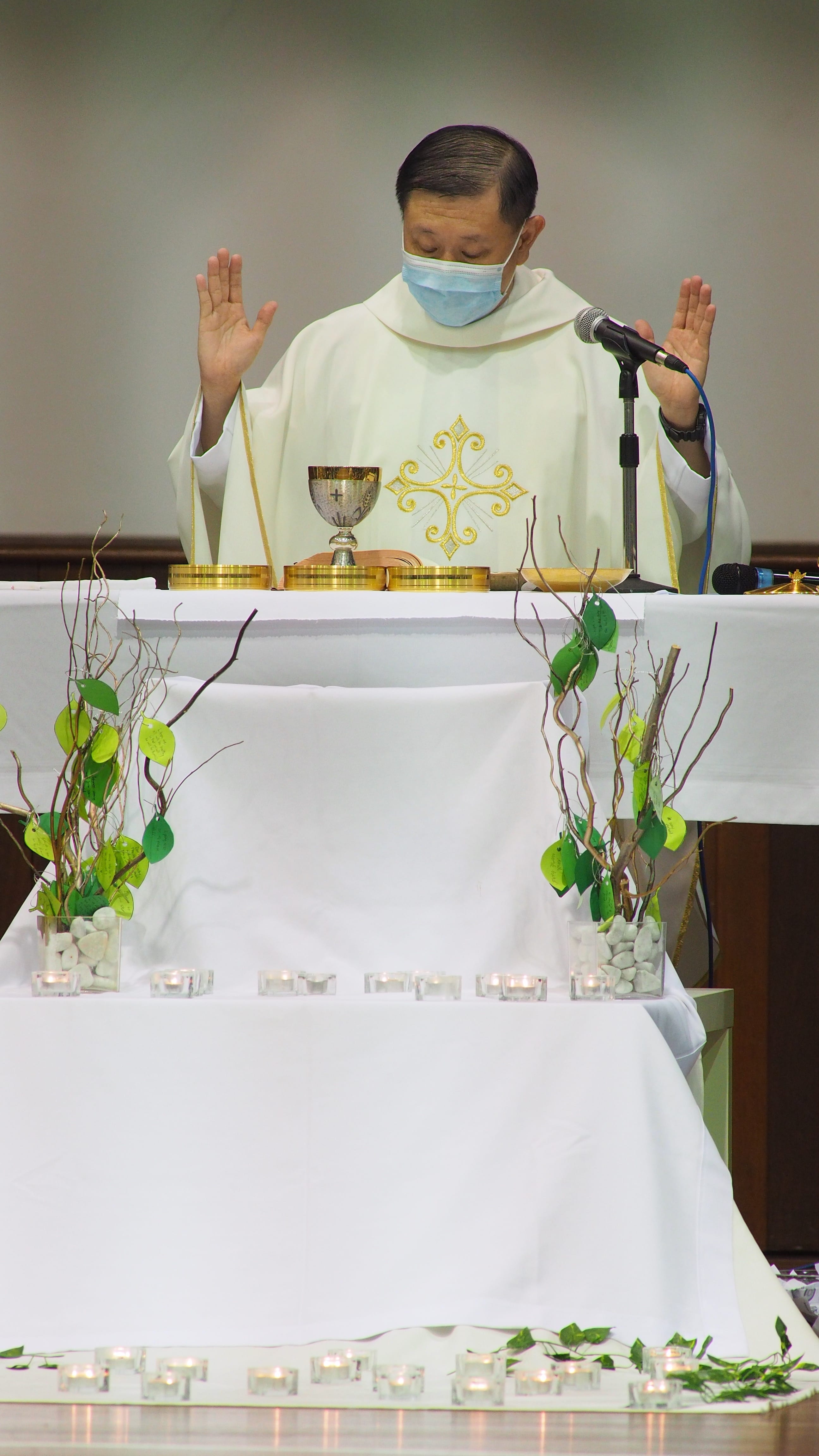 A priest in white vestments and a face mask raises both hands at an altar with a chalice, candles, and green branch decorations.
