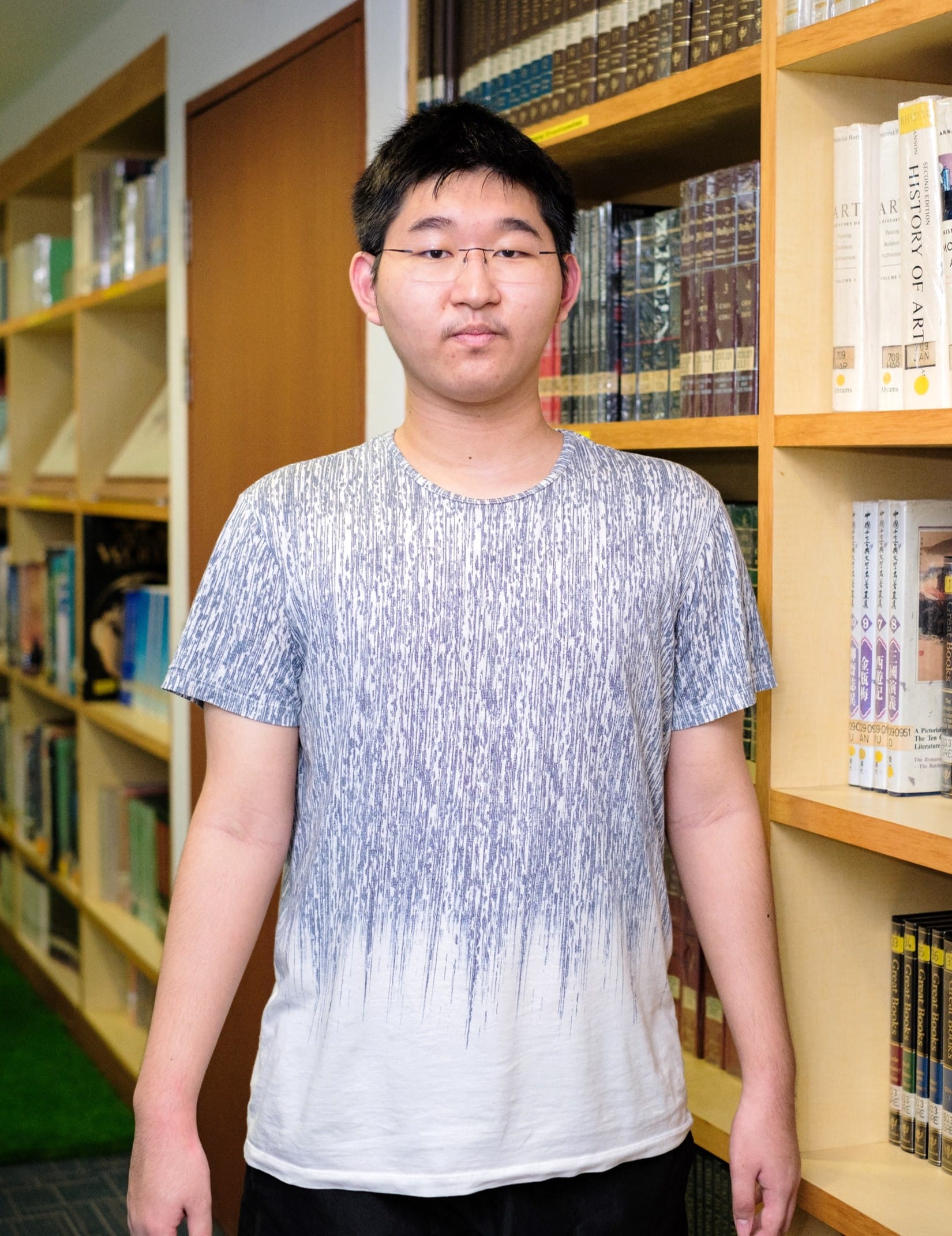 Young man with glasses and a patterned gray t-shirt standing in front of wooden library shelves filled with books.