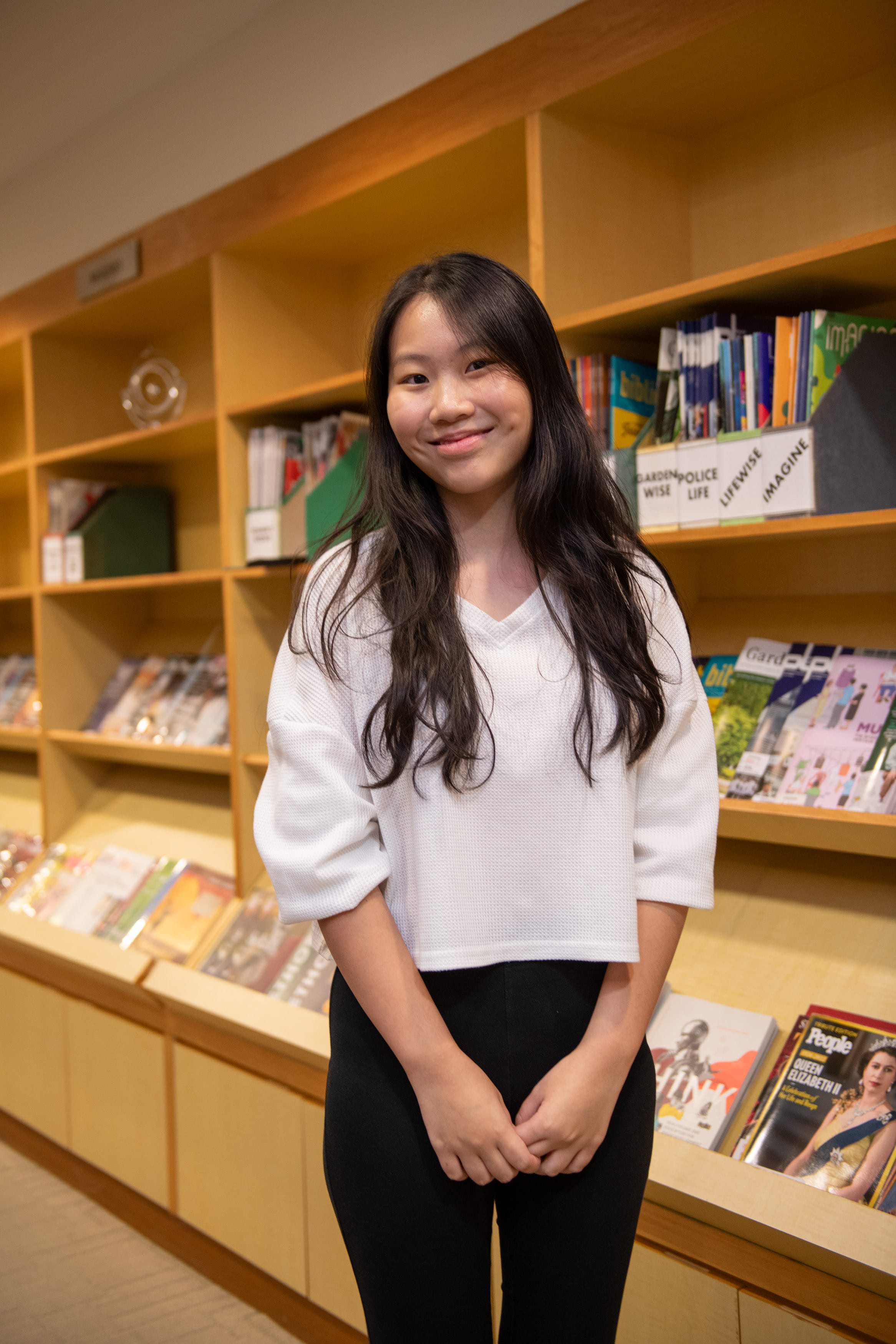 Young woman with long dark hair in a white top and black pants smiling in front of wooden library shelves with magazines.