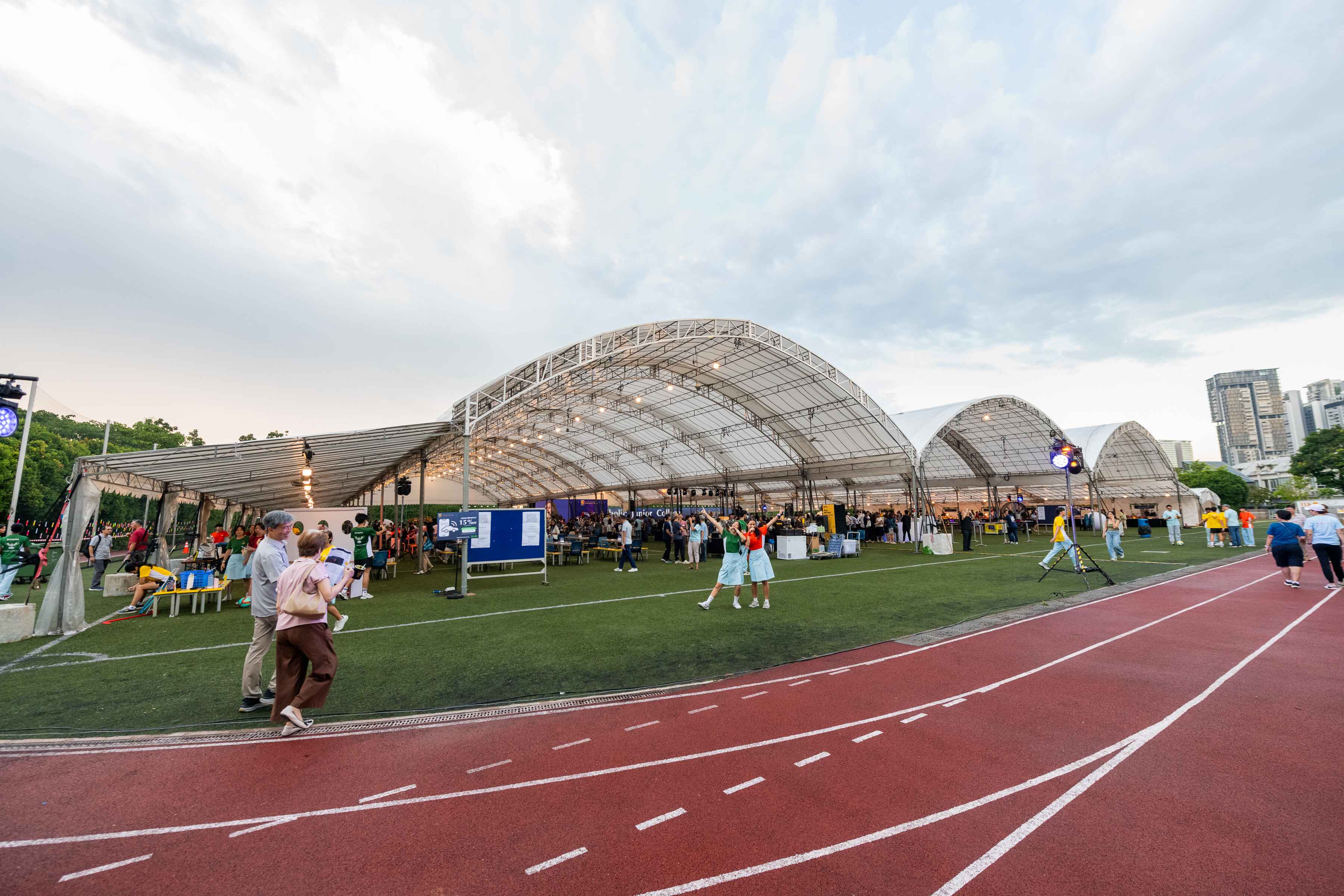 Outdoor event on a green field inside large arched tents, bordered by a red running track, with crowds of people.