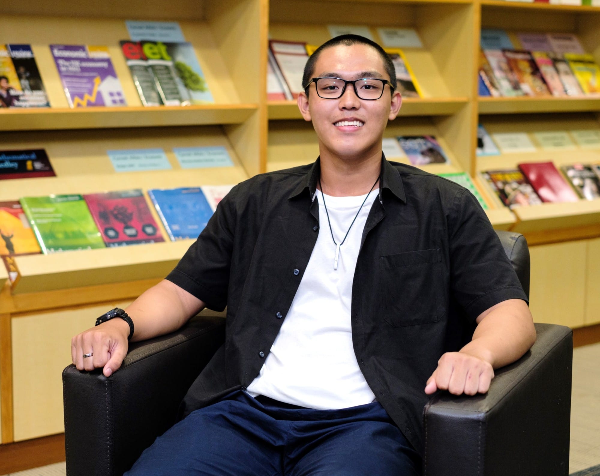 Young man with glasses and short hair smiles while seated in a black chair in front of magazine-filled shelves.