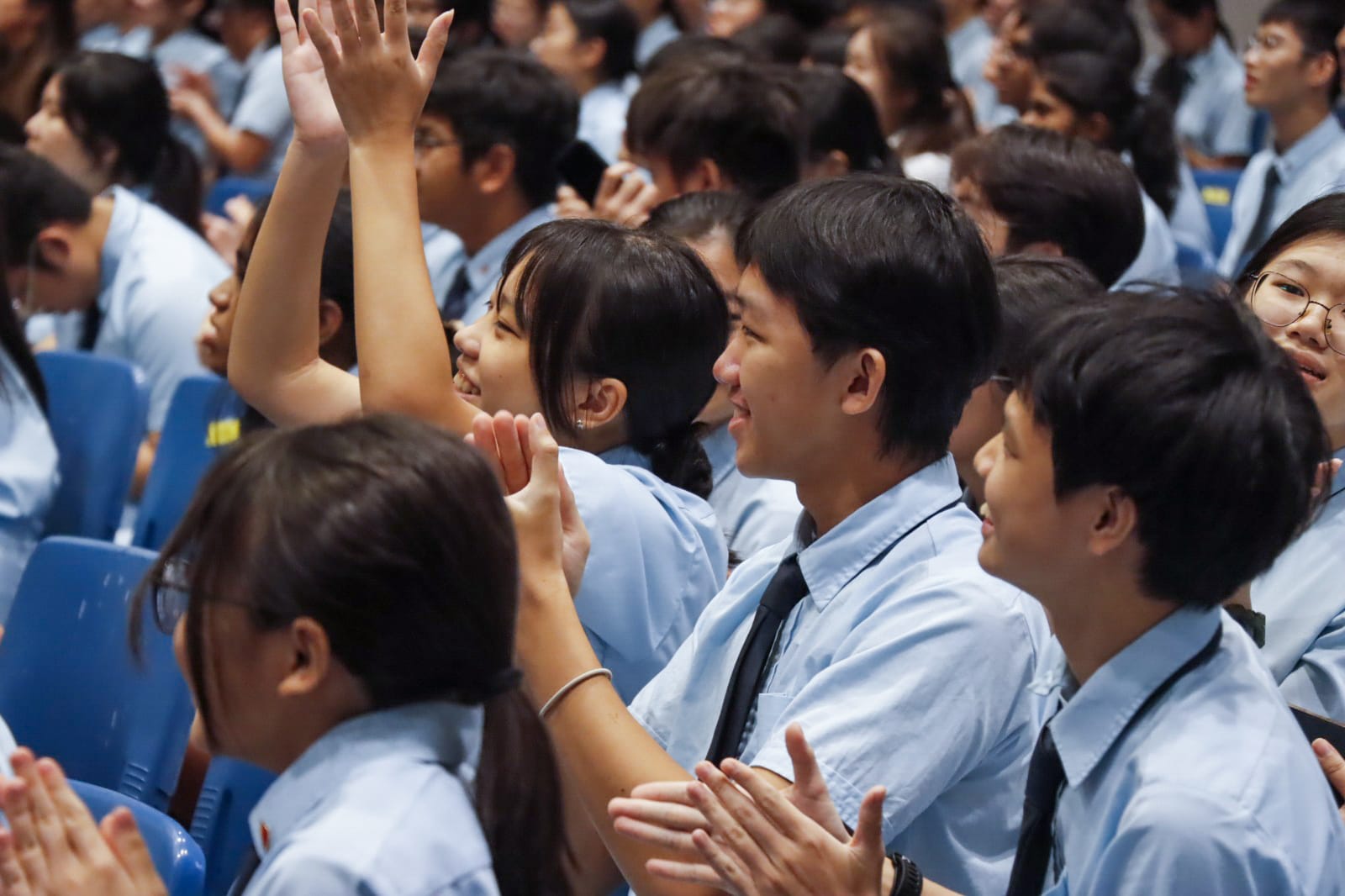 A crowd of students in light blue uniforms clapping and raising hands, seated in blue chairs at a school assembly.
