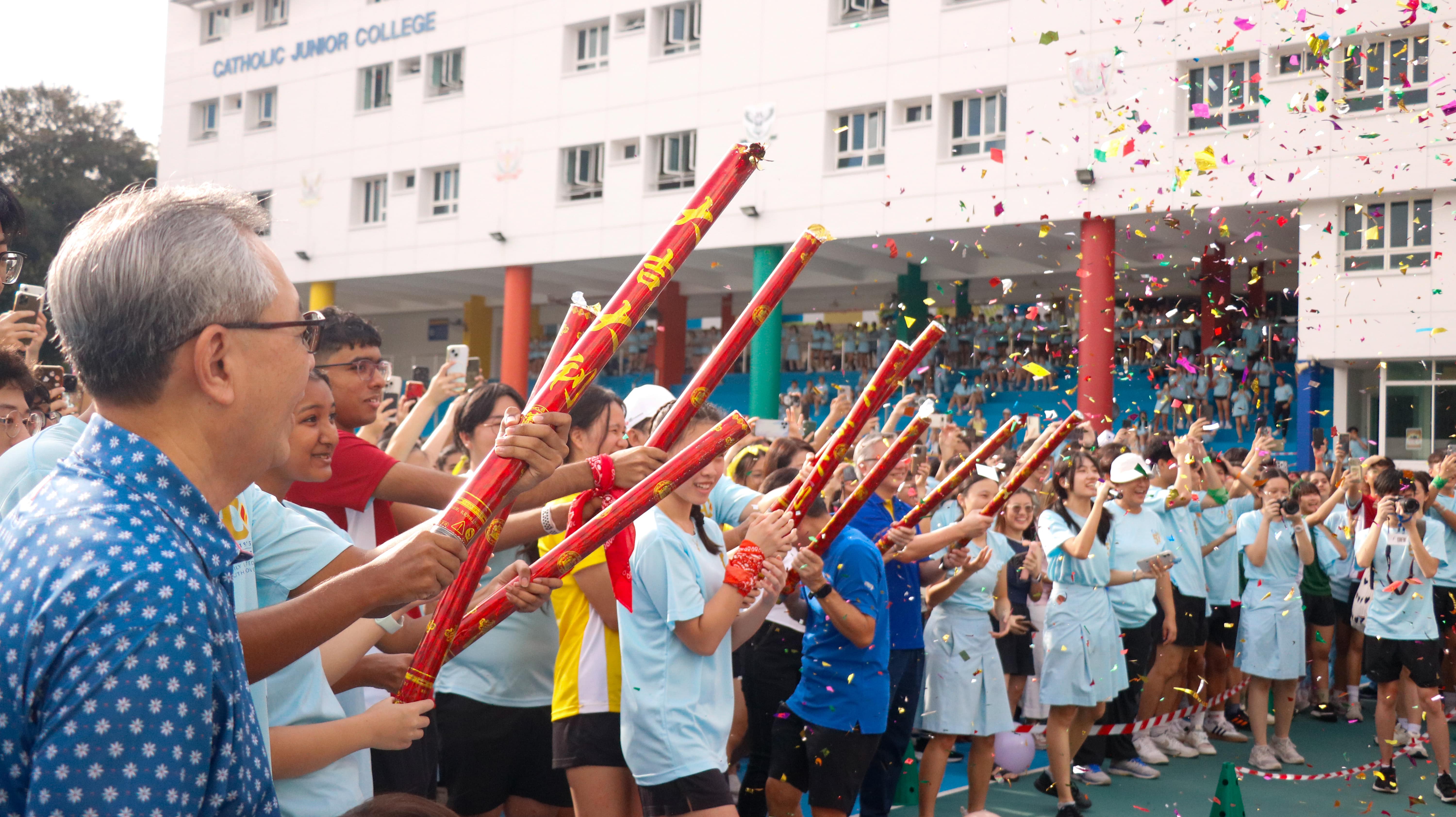 Students and staff at Catholic Junior College hold red confetti cannons as colorful confetti fills the air.