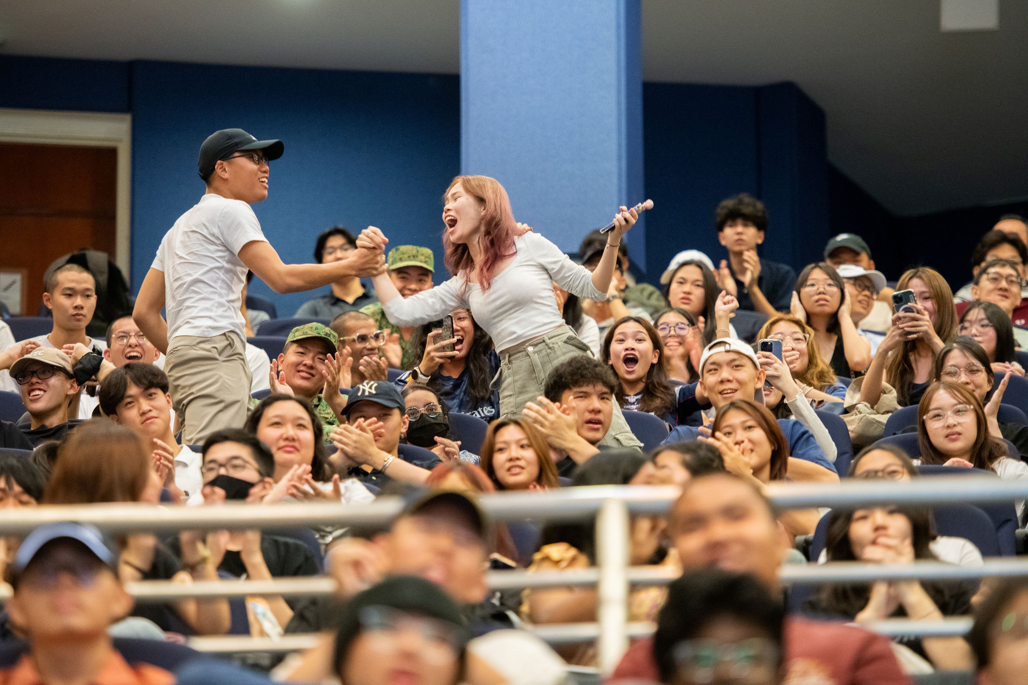 Two young people stand cheering in a packed auditorium crowd, with excited audience members clapping around them.