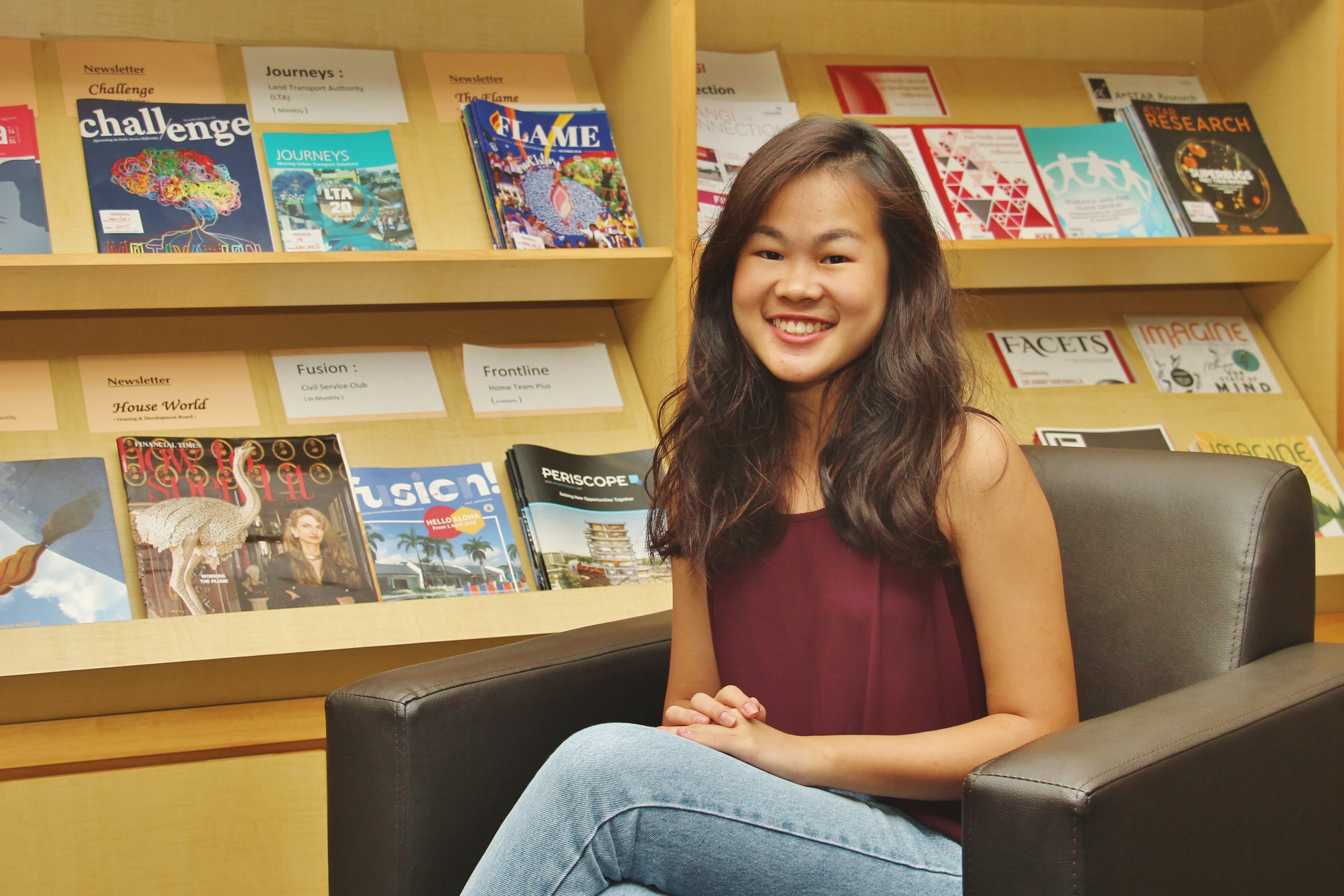 Smiling woman with long hair sits in a black armchair in front of shelves filled with magazines.