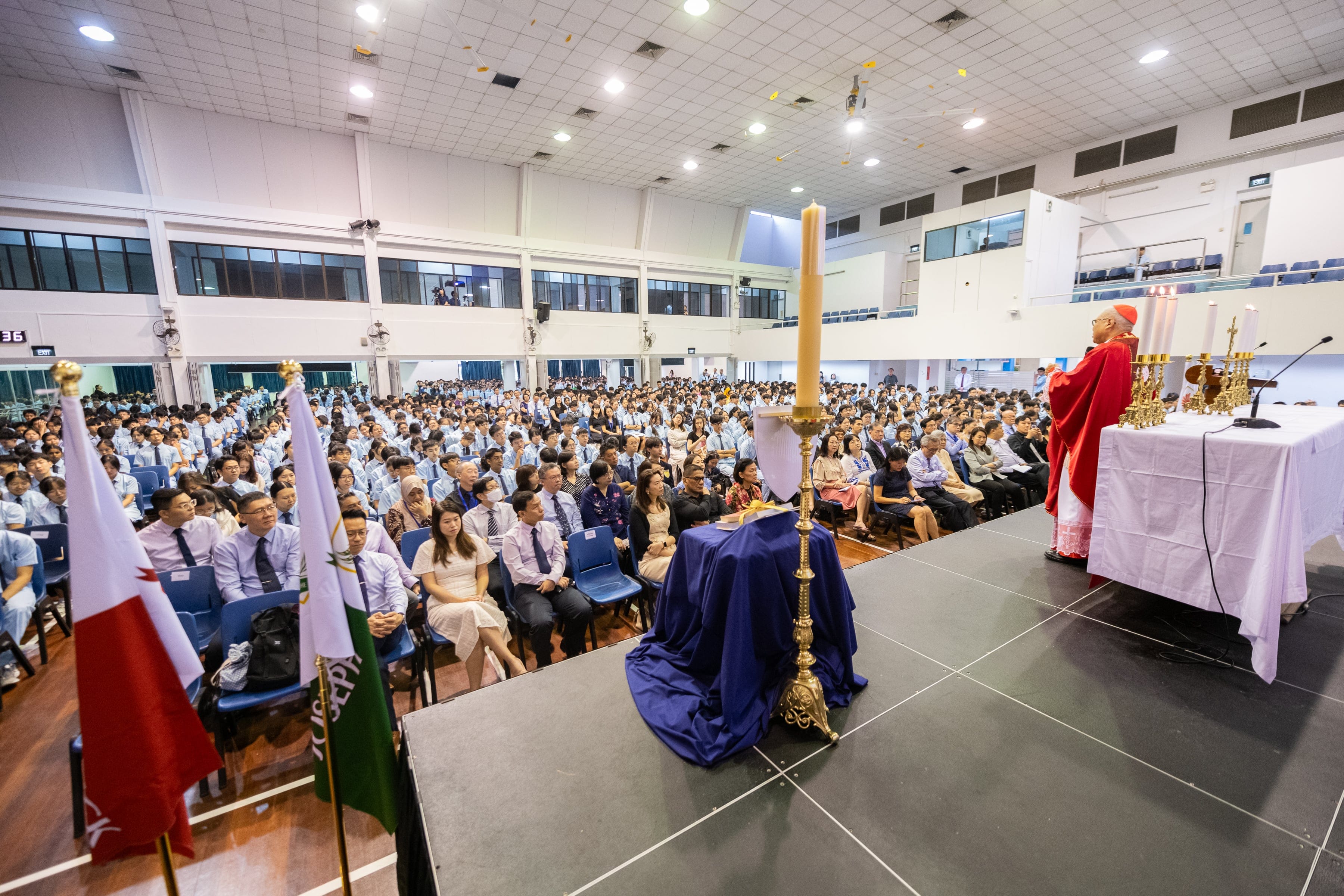 A Cardinal in red robes leads a Catholic Mass on stage before a large seated crowd in a school hall.