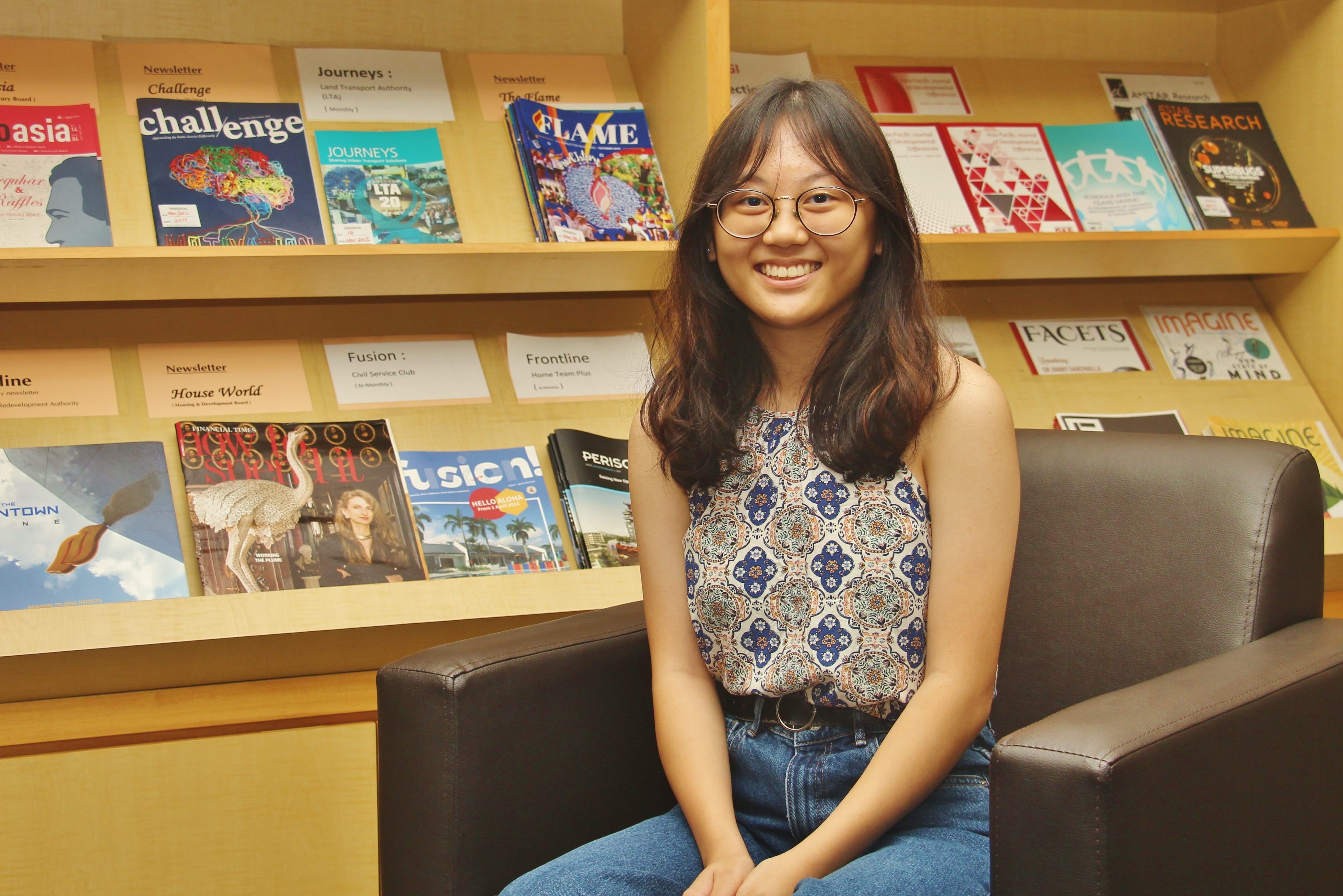 Smiling woman with glasses sits in an armchair in front of shelves filled with magazines and newsletters.