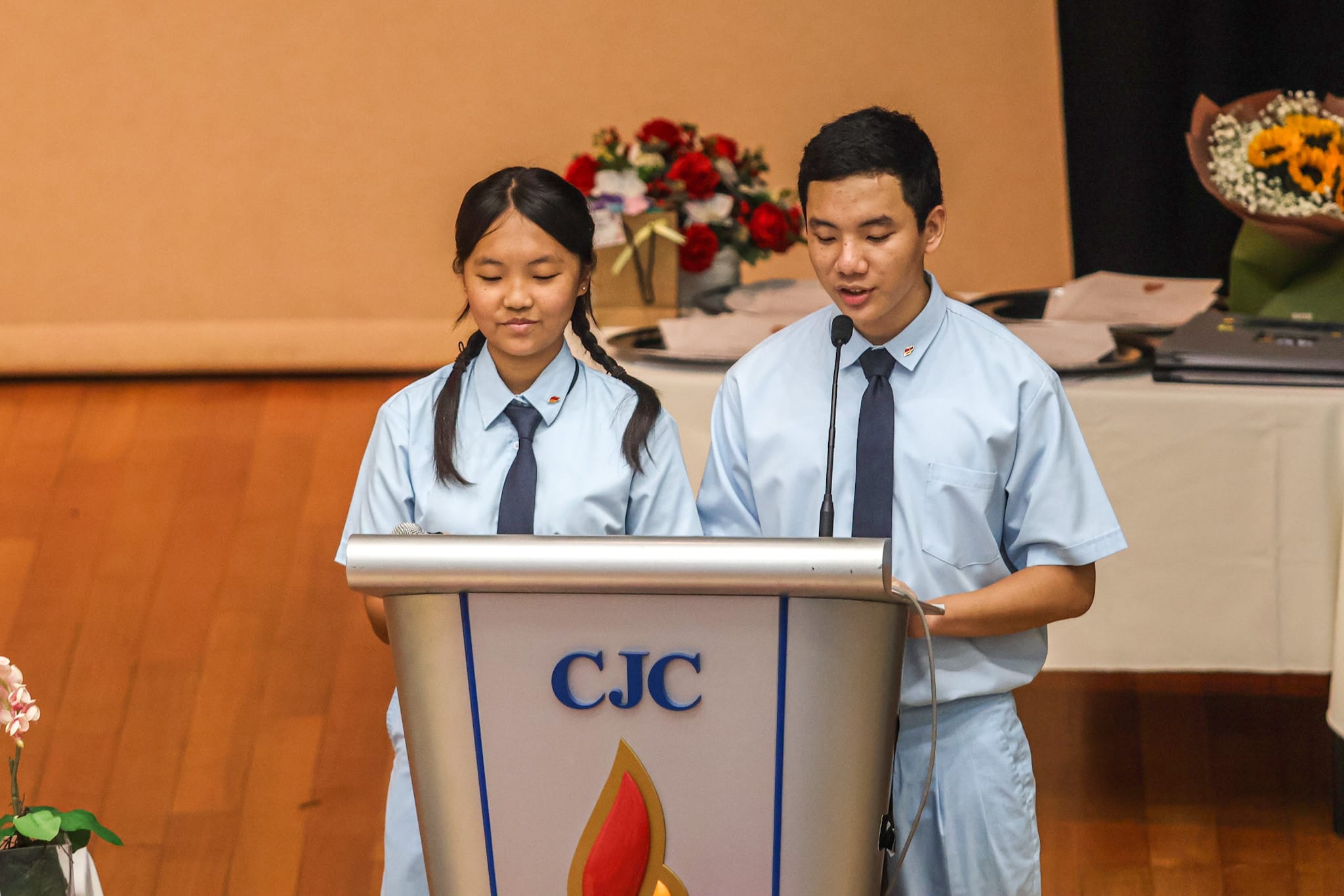 Two students in light blue uniforms with ties stand at a CJC podium with a microphone on stage.