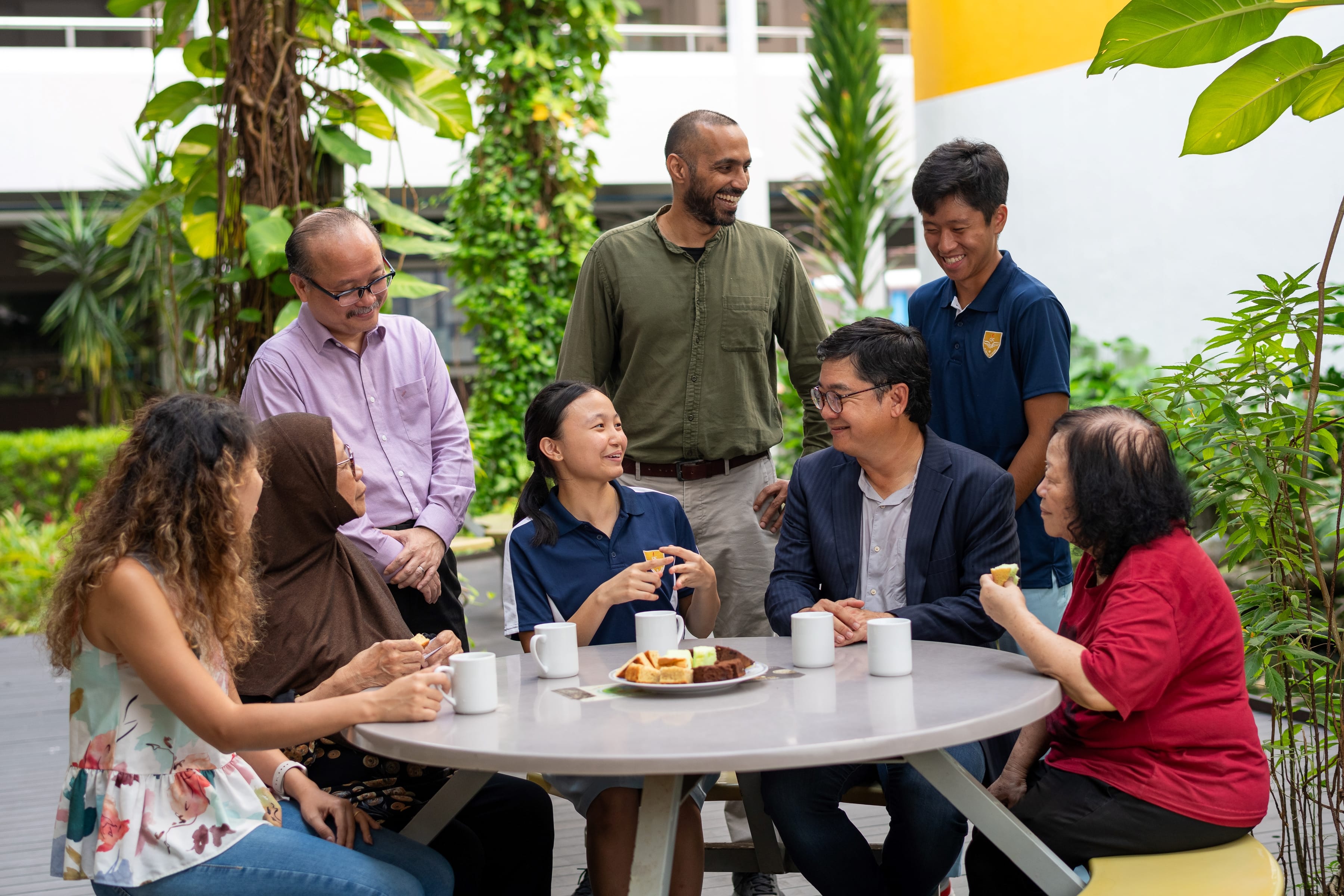 Eight people chat and smile around a table with mugs and snacks in a leafy outdoor courtyard.