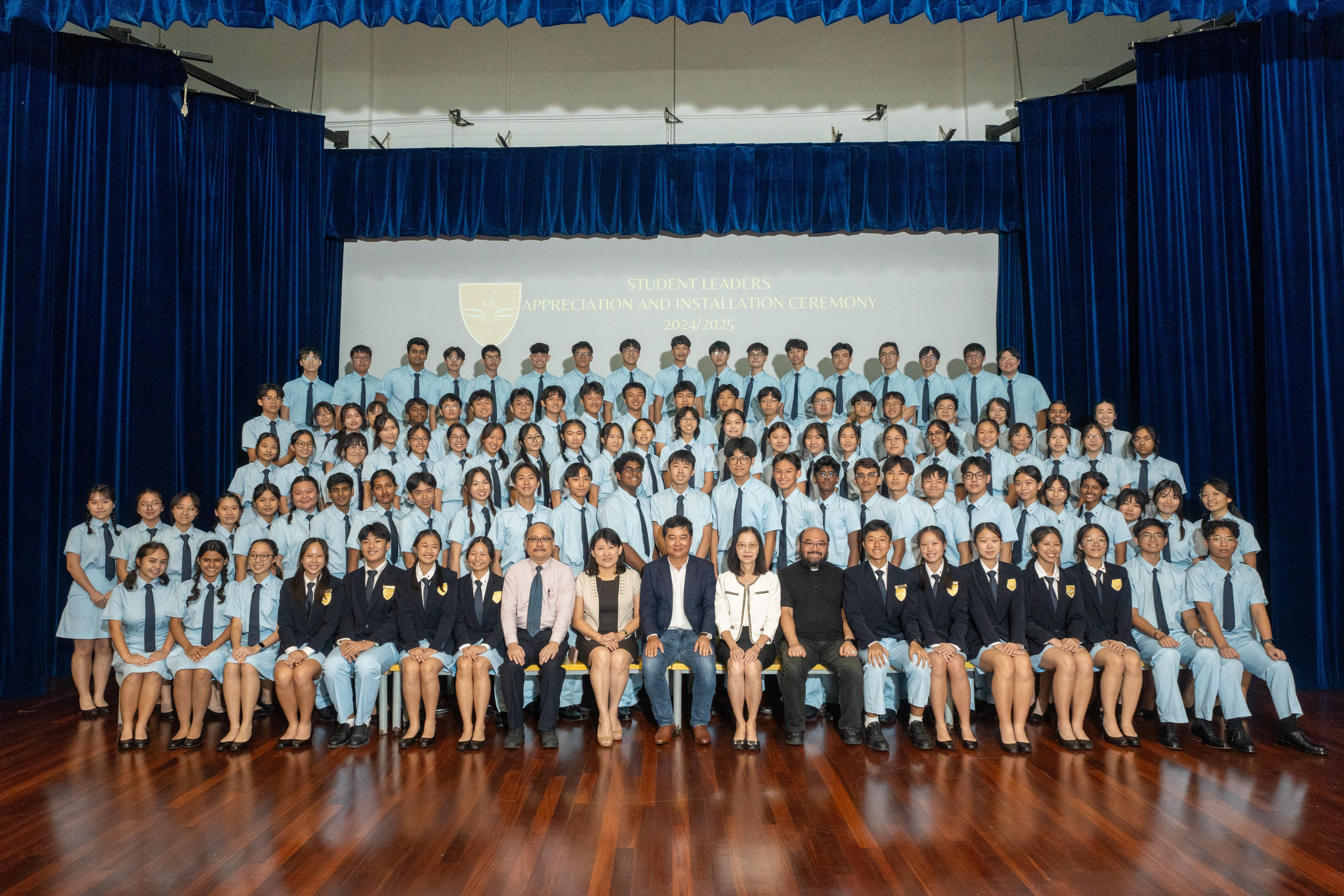 Large group of students in matching uniforms posed on a stage with blue curtains, with staff seated in front.
