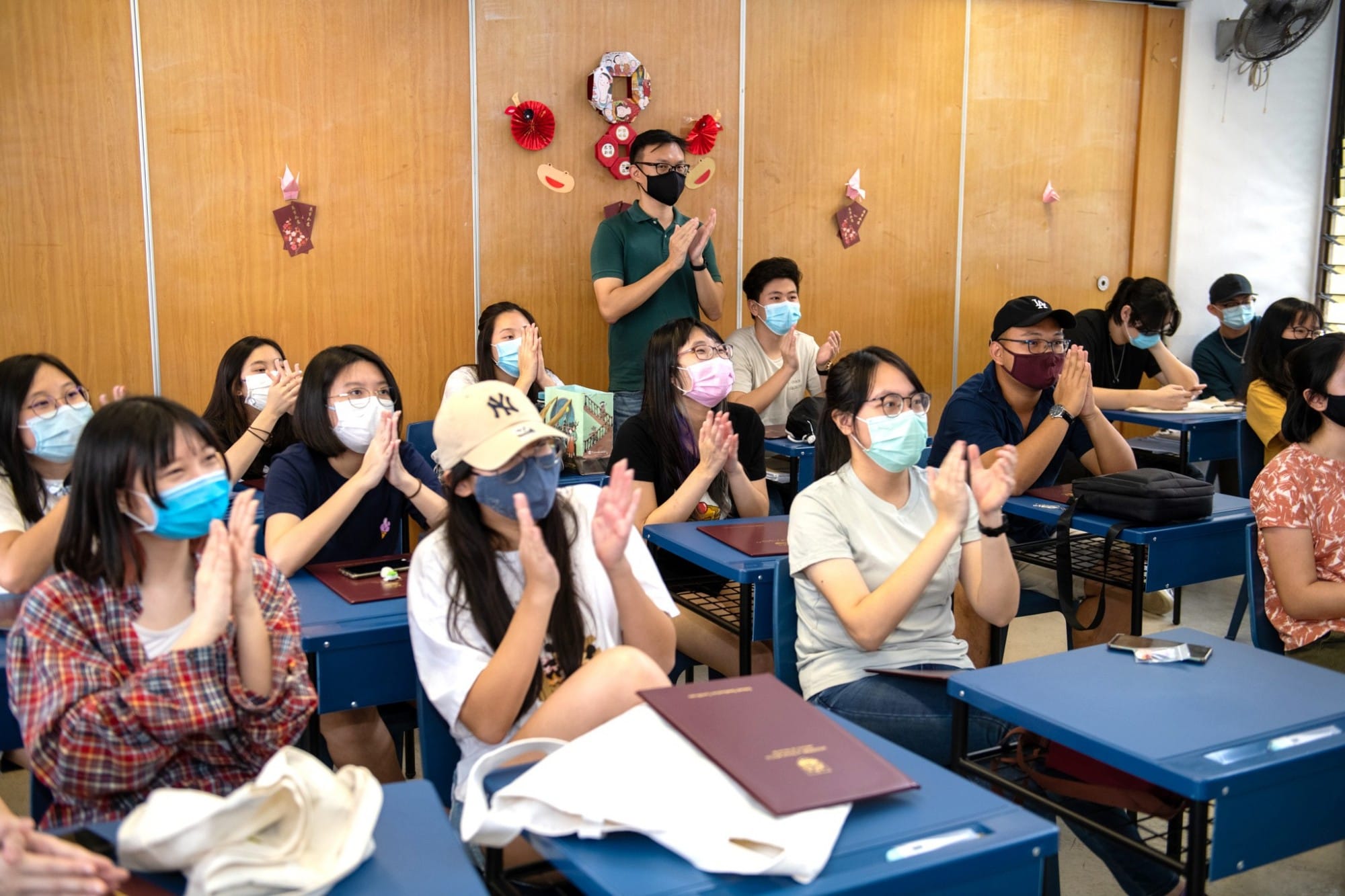 Students wearing face masks sit at blue desks in a decorated classroom, clapping together with a standing teacher.