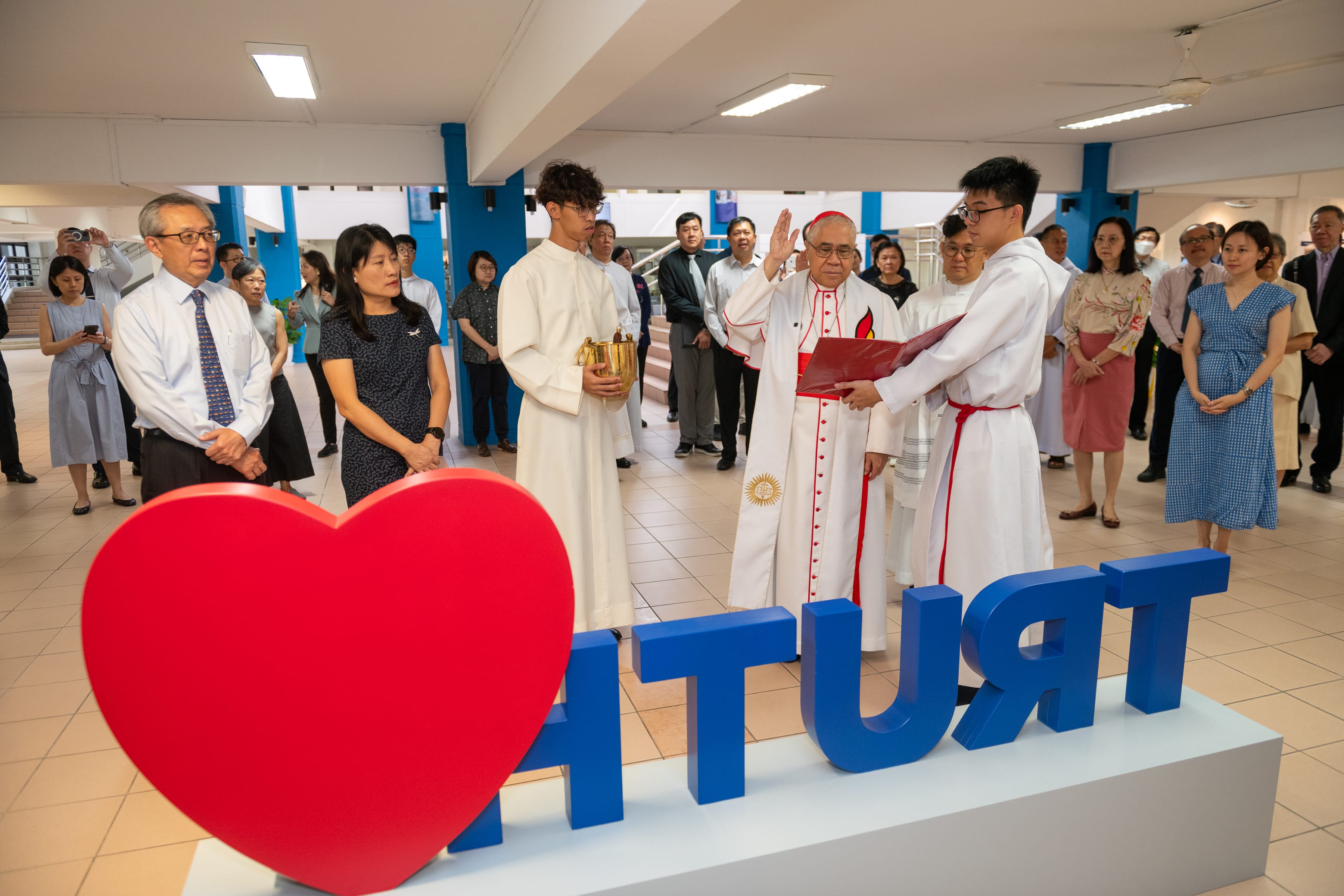 Clergy in white robes lead a ceremony as people watch, behind a large red heart and blue letters.