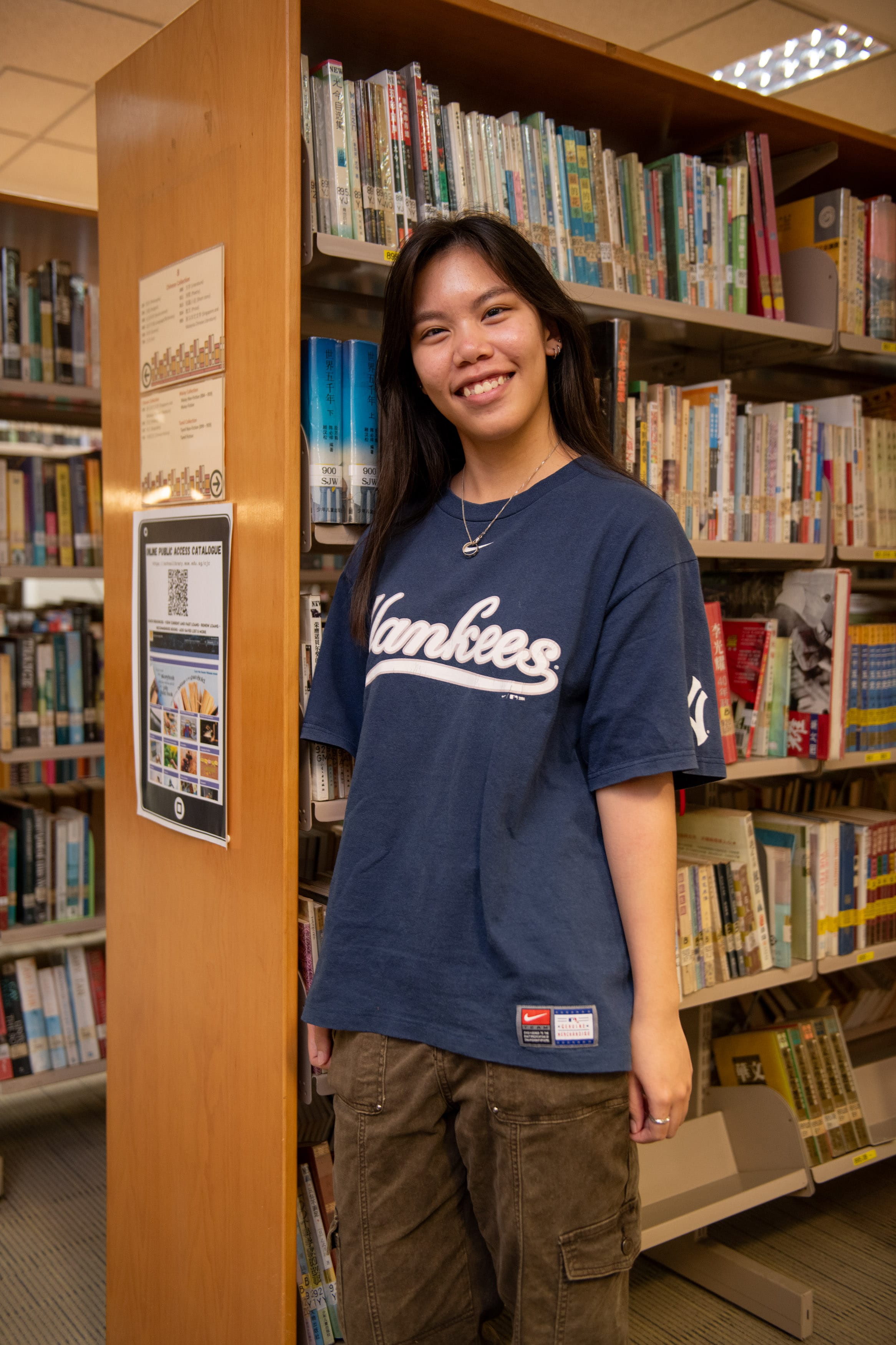 Young woman smiling in a library, wearing a navy Yankees shirt and olive cargo pants, standing beside shelves of books.